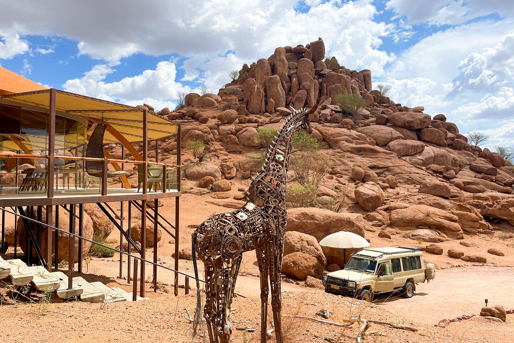 A modern safari lodge with a shaded deck sits among large red rock formations in a desert landscape. In the foreground stands a tall giraffe sculpture made of metal parts, while a safari vehicle with a pop-up roof is parked nearby under a bright, partly cloudy sky.