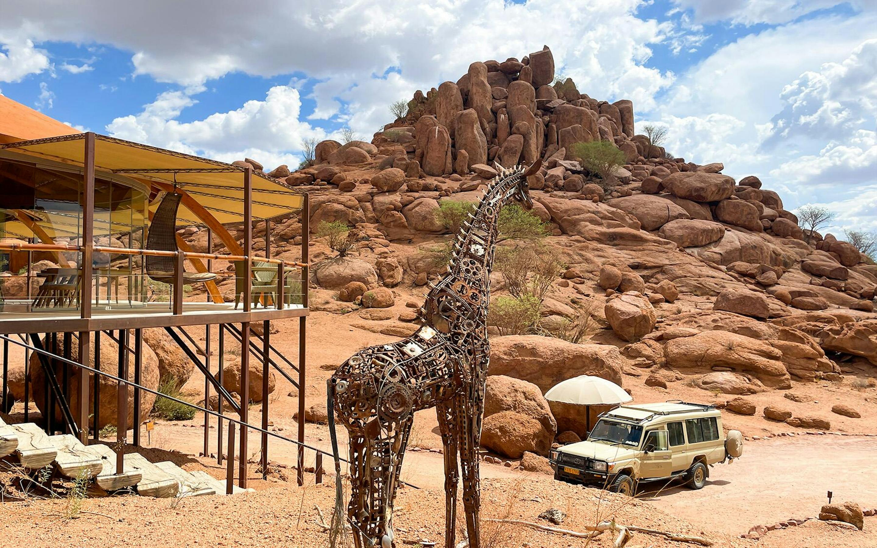 A modern safari lodge with a shaded deck sits among large red rock formations in a desert landscape. In the foreground stands a tall giraffe sculpture made of metal parts, while a safari vehicle with a pop-up roof is parked nearby under a bright, partly cloudy sky.
