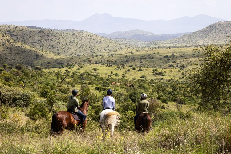 Three people on horseback look out over a vast plain in Kenya.