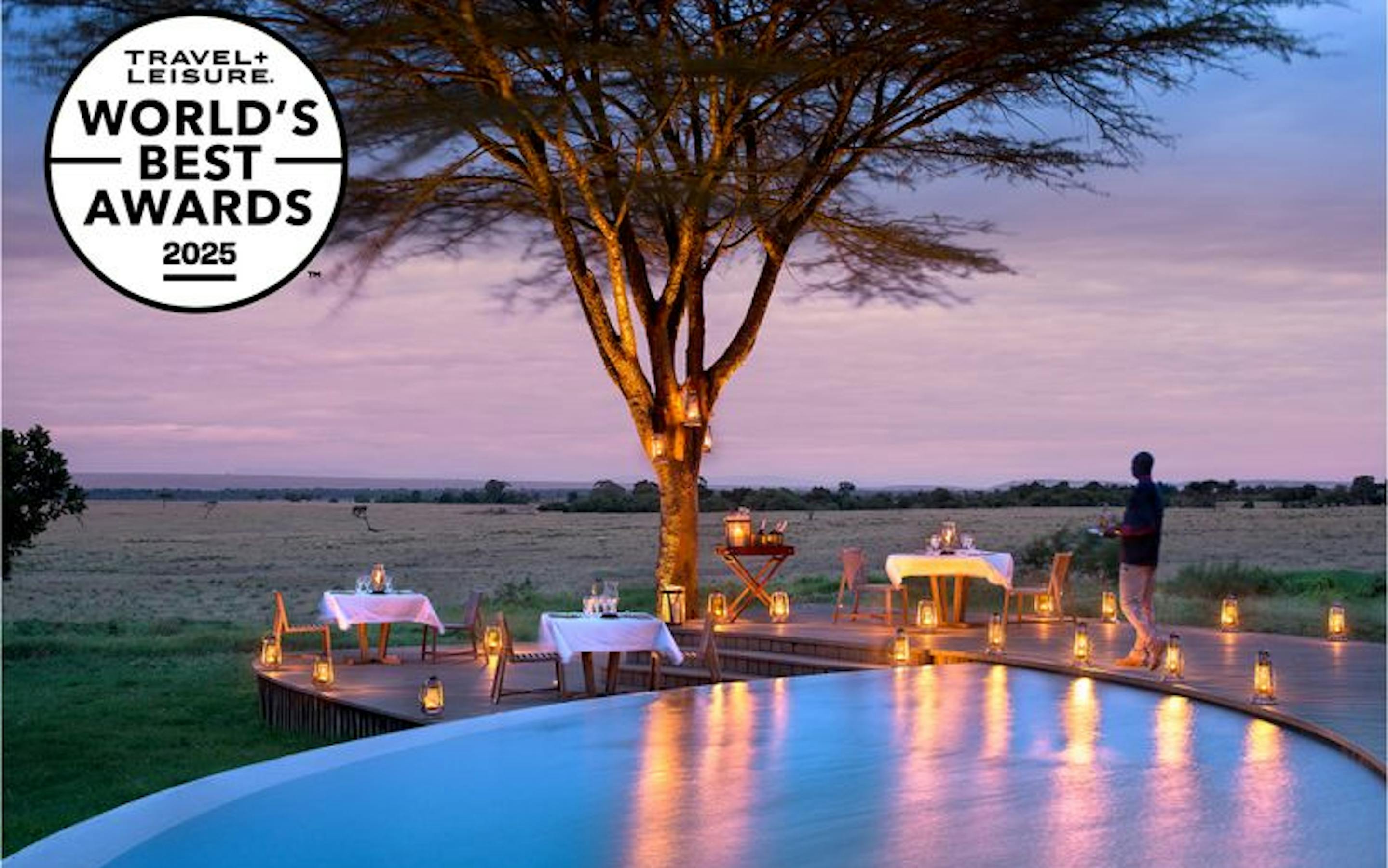 An infinity pool overlooks an open savanna at sunset, where lantern-lit dining tables are set beneath a large tree. A circular badge in the corner reads “Travel + Leisure World’s Best Awards 2025.”