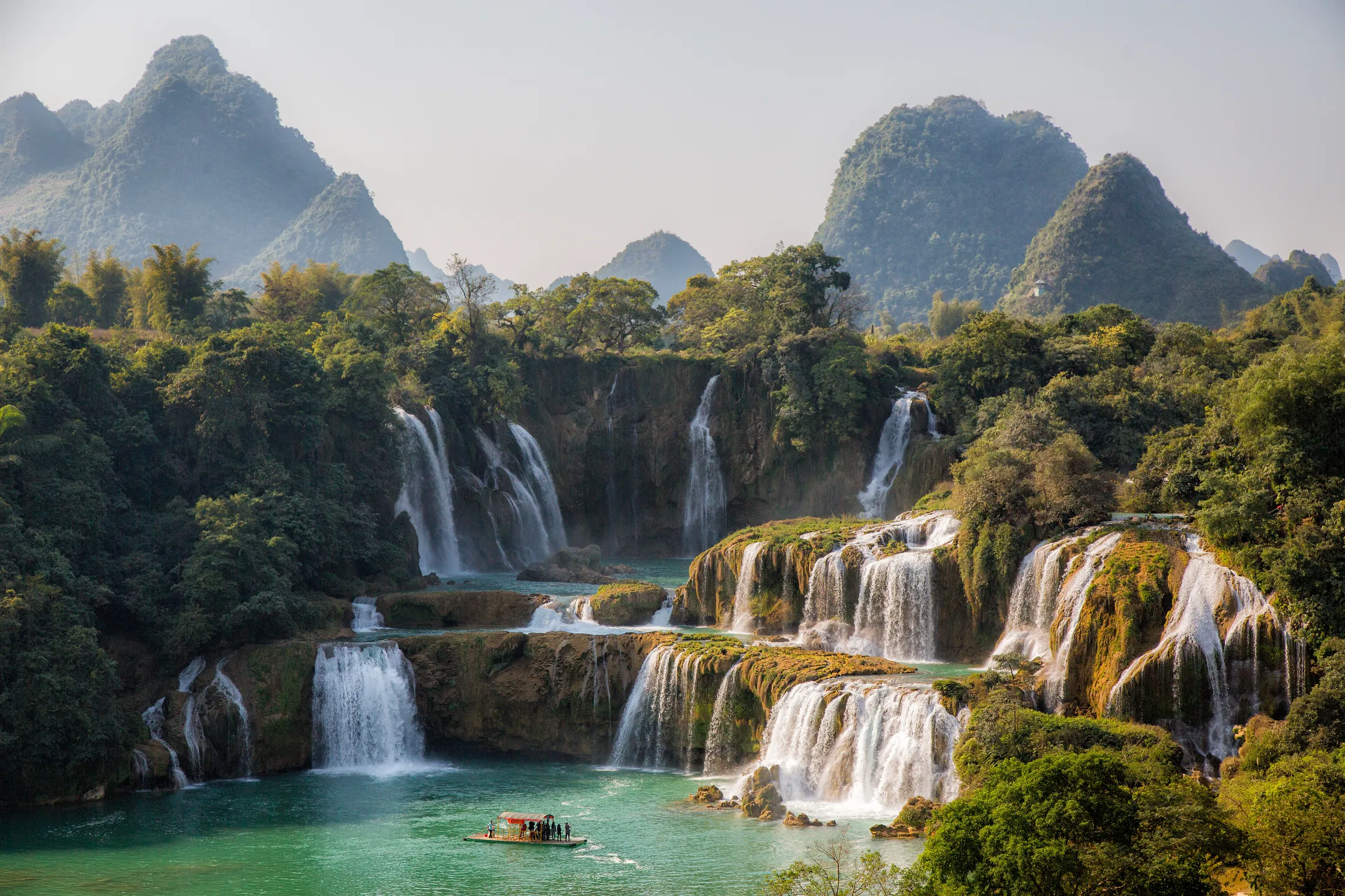 Multi-tiered waterfalls cascade into turquoise pools surrounded by lush green forest, with mist rising against a backdrop of rounded limestone mountains. A small boat with passengers floats on the water below the falls.