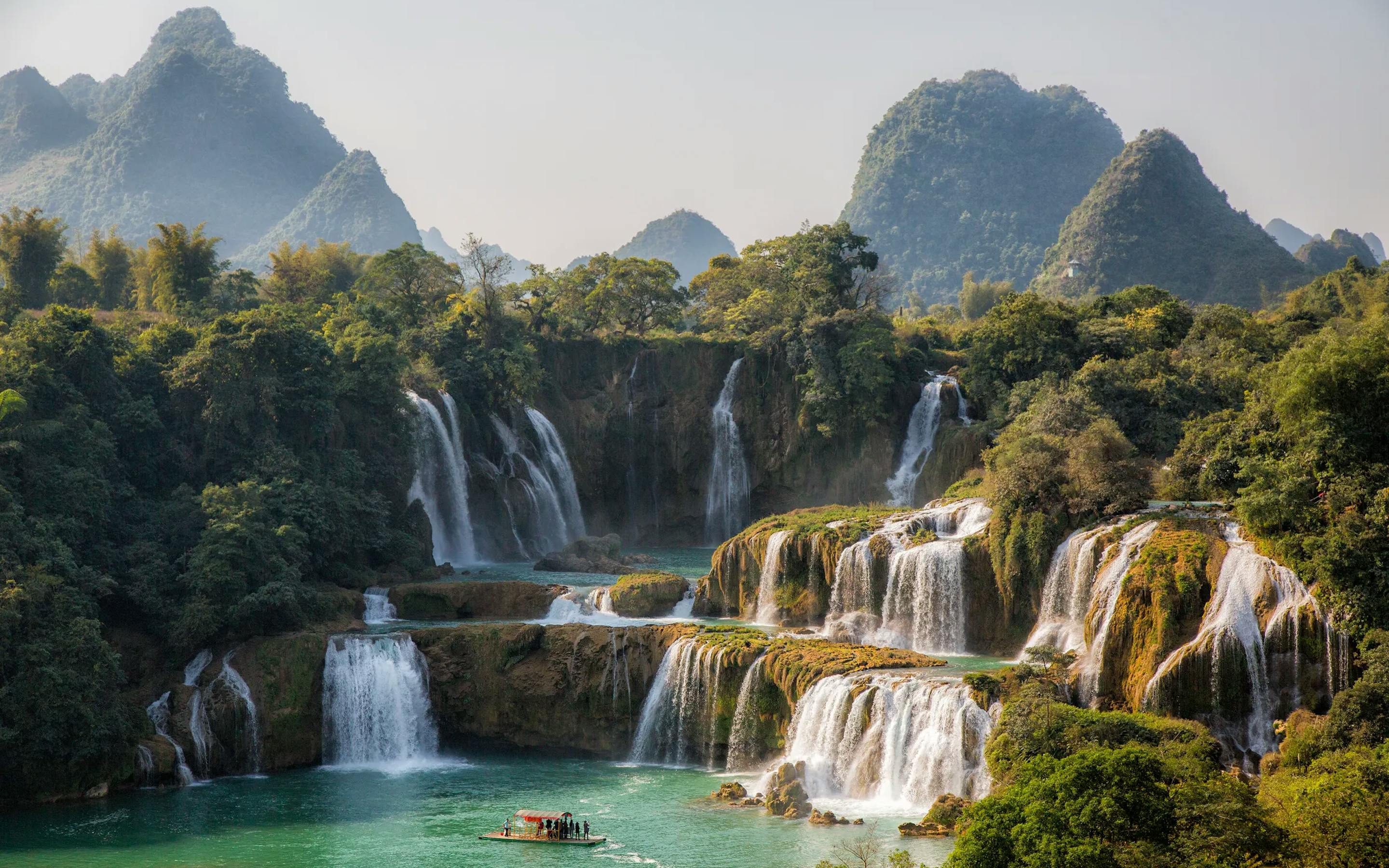 Multi-tiered waterfalls cascade into turquoise pools surrounded by lush green forest, with mist rising against a backdrop of rounded limestone mountains. A small boat with passengers floats on the water below the falls.