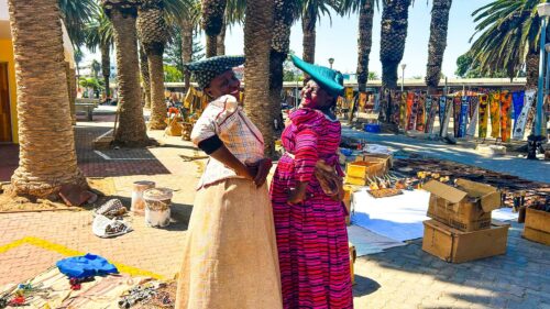 Two women in colorful traditional dresses and headwraps stand facing each other at an outdoor market lined with palm trees, with handmade goods displayed on the ground and in nearby stalls.