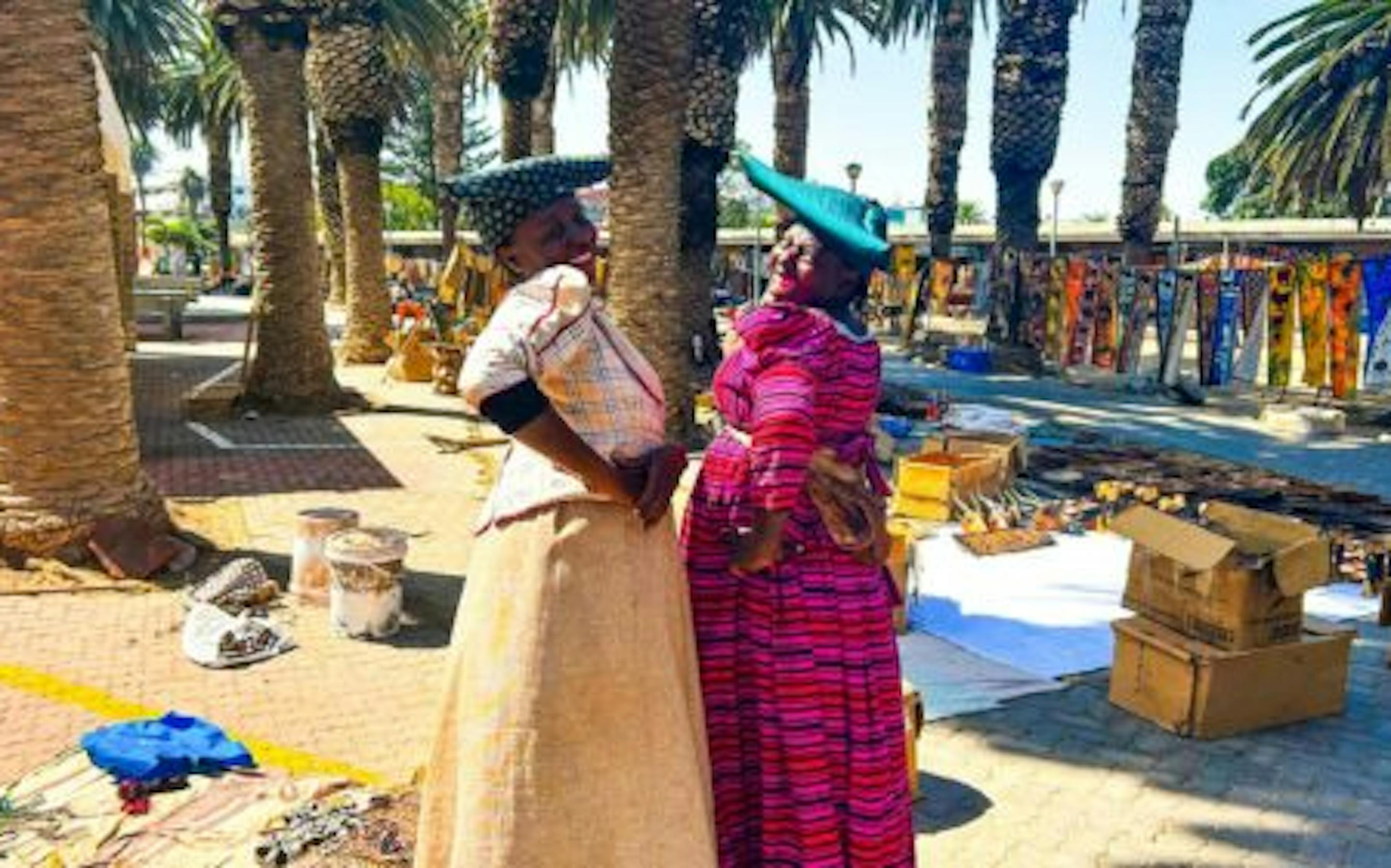 Two women in colorful traditional dresses and headwraps stand facing each other at an outdoor market lined with palm trees, with handmade goods displayed on the ground and in nearby stalls.
