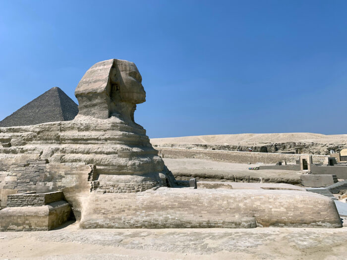 The Great Sphinx of Giza stands in the desert under a clear blue sky, with one of the pyramids visible in the background.