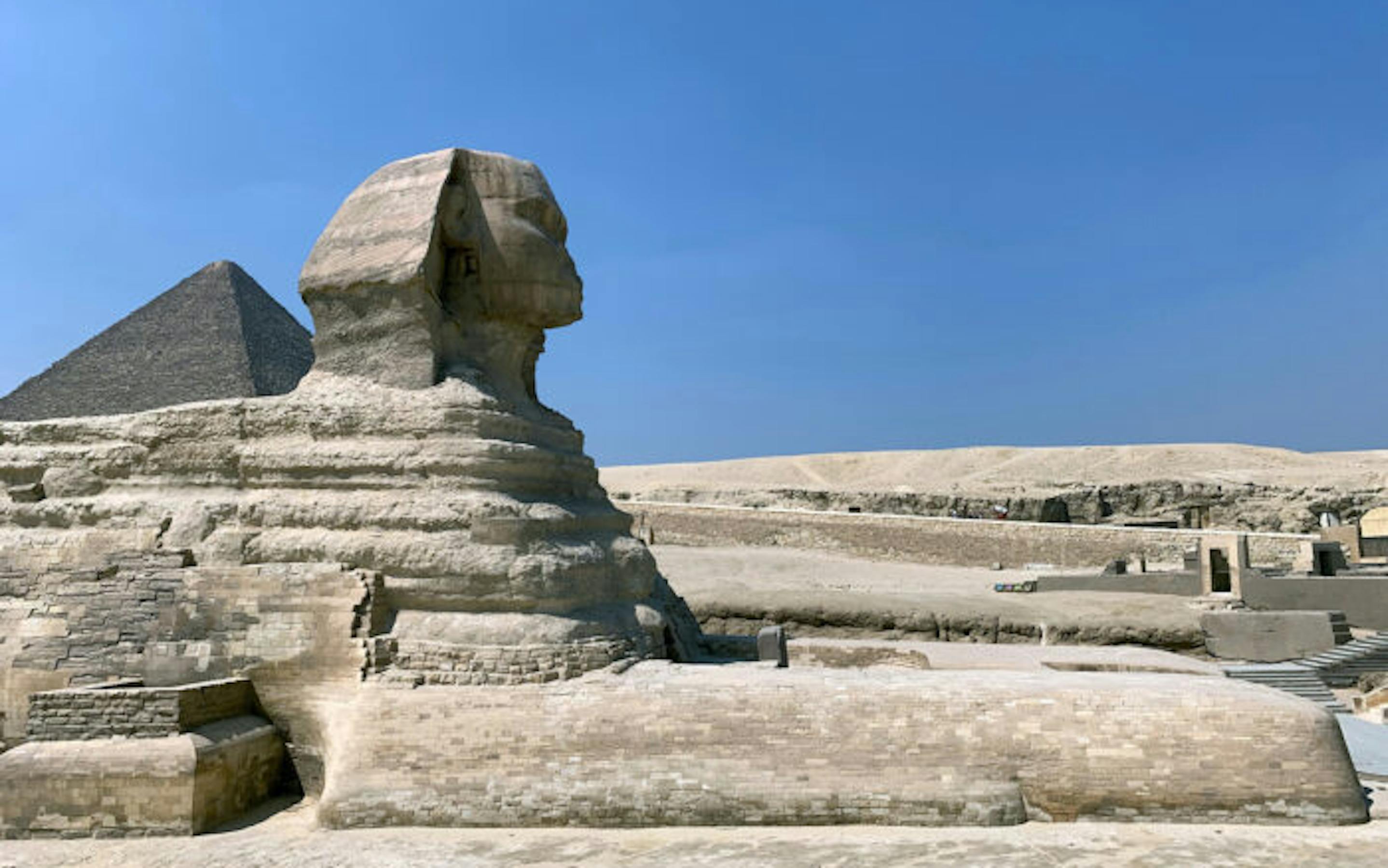 The Great Sphinx of Giza stands in the desert under a clear blue sky, with one of the pyramids visible in the background.
