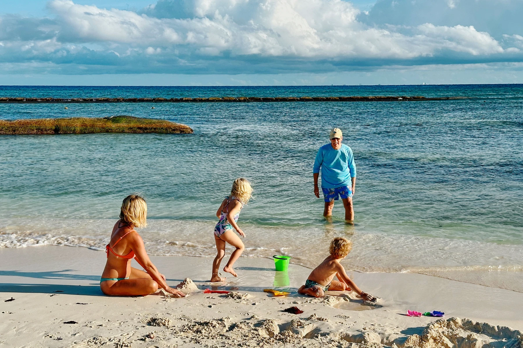 A family plays on a sandy beach by clear turquoise water, with two young children building sandcastles near the shore while an adult stands in the shallow ocean under a bright, partly cloudy sky.