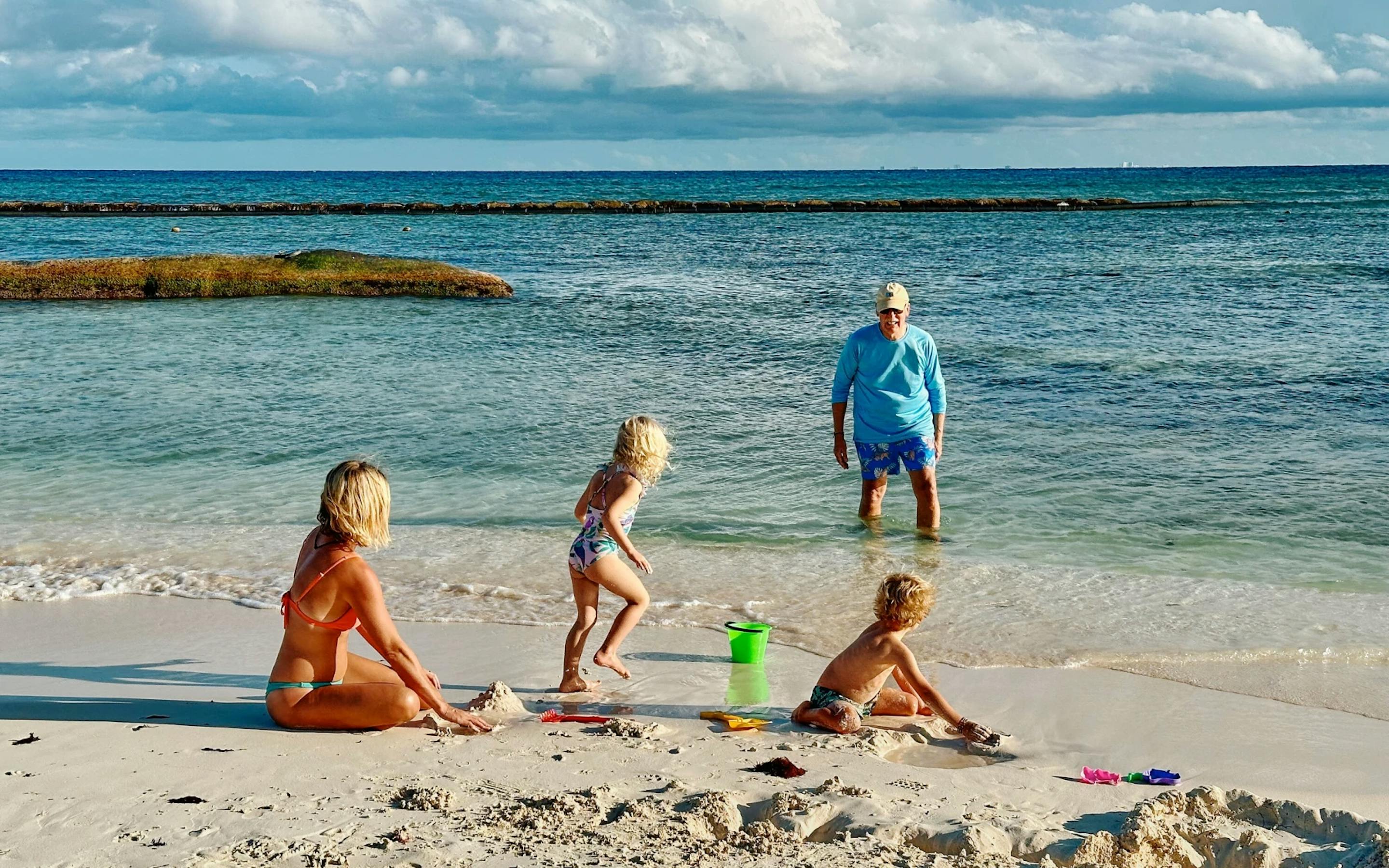 A family plays on a sandy beach by clear turquoise water, with two young children building sandcastles near the shore while an adult stands in the shallow ocean under a bright, partly cloudy sky.