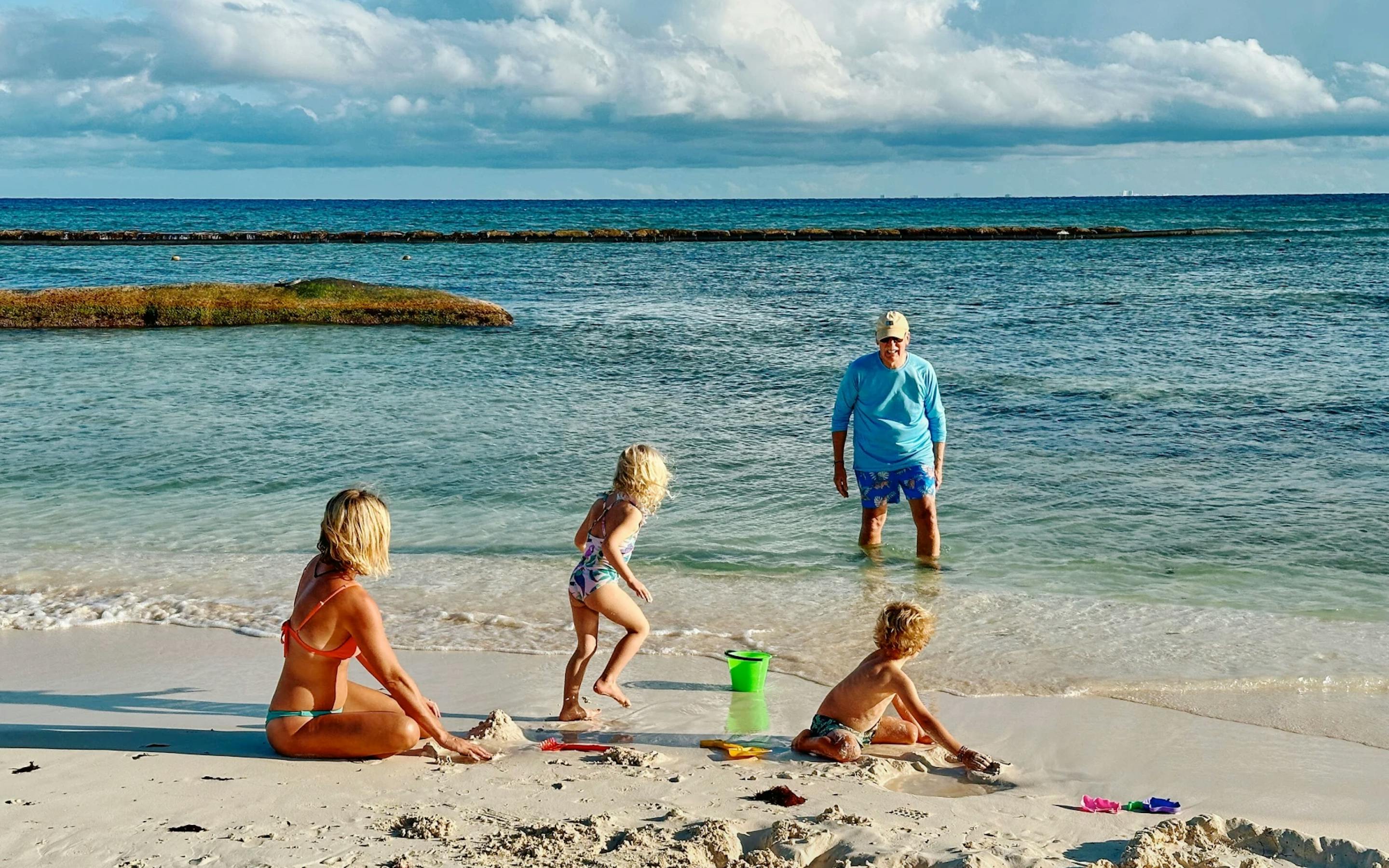 A family plays on a sandy beach by clear turquoise water, with two young children building sandcastles near the shore while an adult stands in the shallow ocean under a bright, partly cloudy sky.
