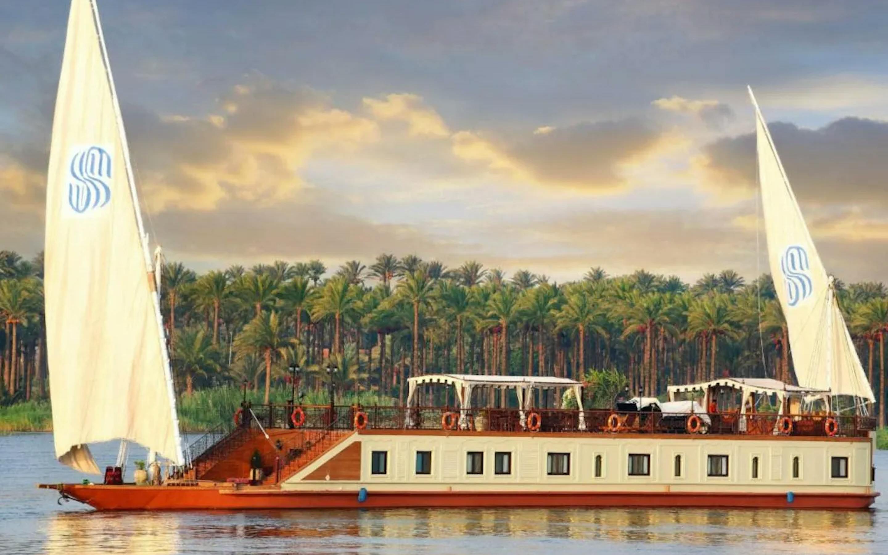 A traditional sailboat with two large white sails glides along a calm river at sunset, with a lush line of palm trees stretching across the shoreline in the background.
