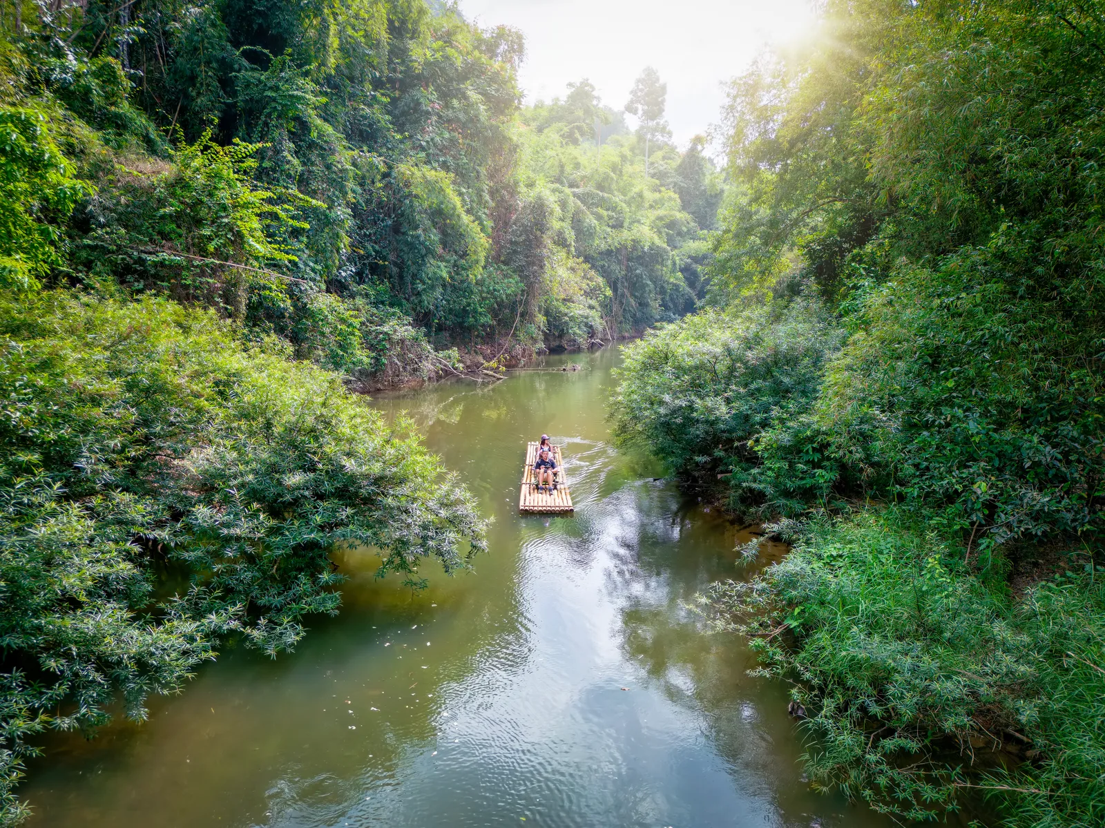 A small bamboo raft carrying two people floats along a calm river surrounded by dense, lush jungle vegetation, with sunlight filtering through the treetops.