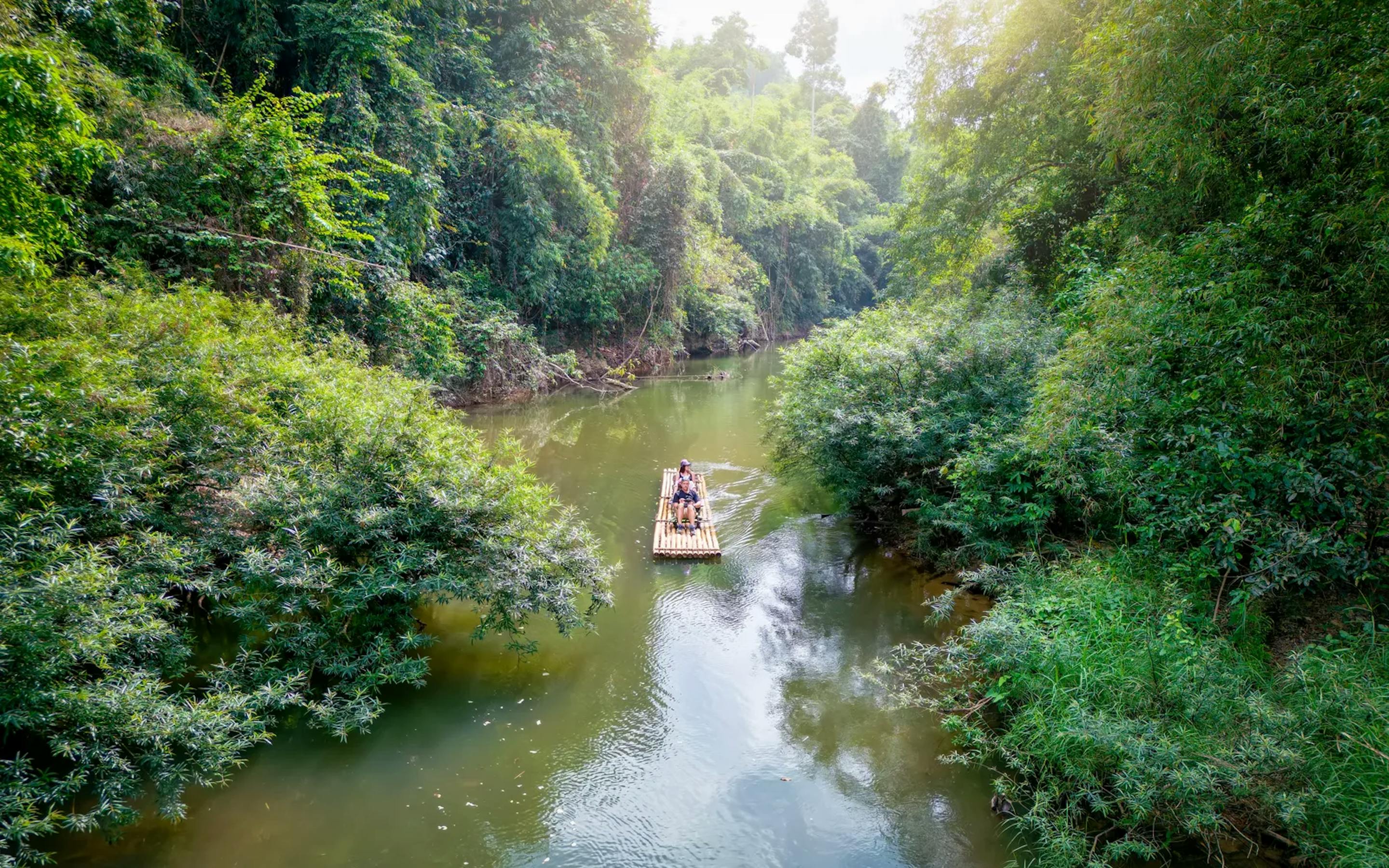 A small bamboo raft carrying two people floats along a calm river surrounded by dense, lush jungle vegetation, with sunlight filtering through the treetops.