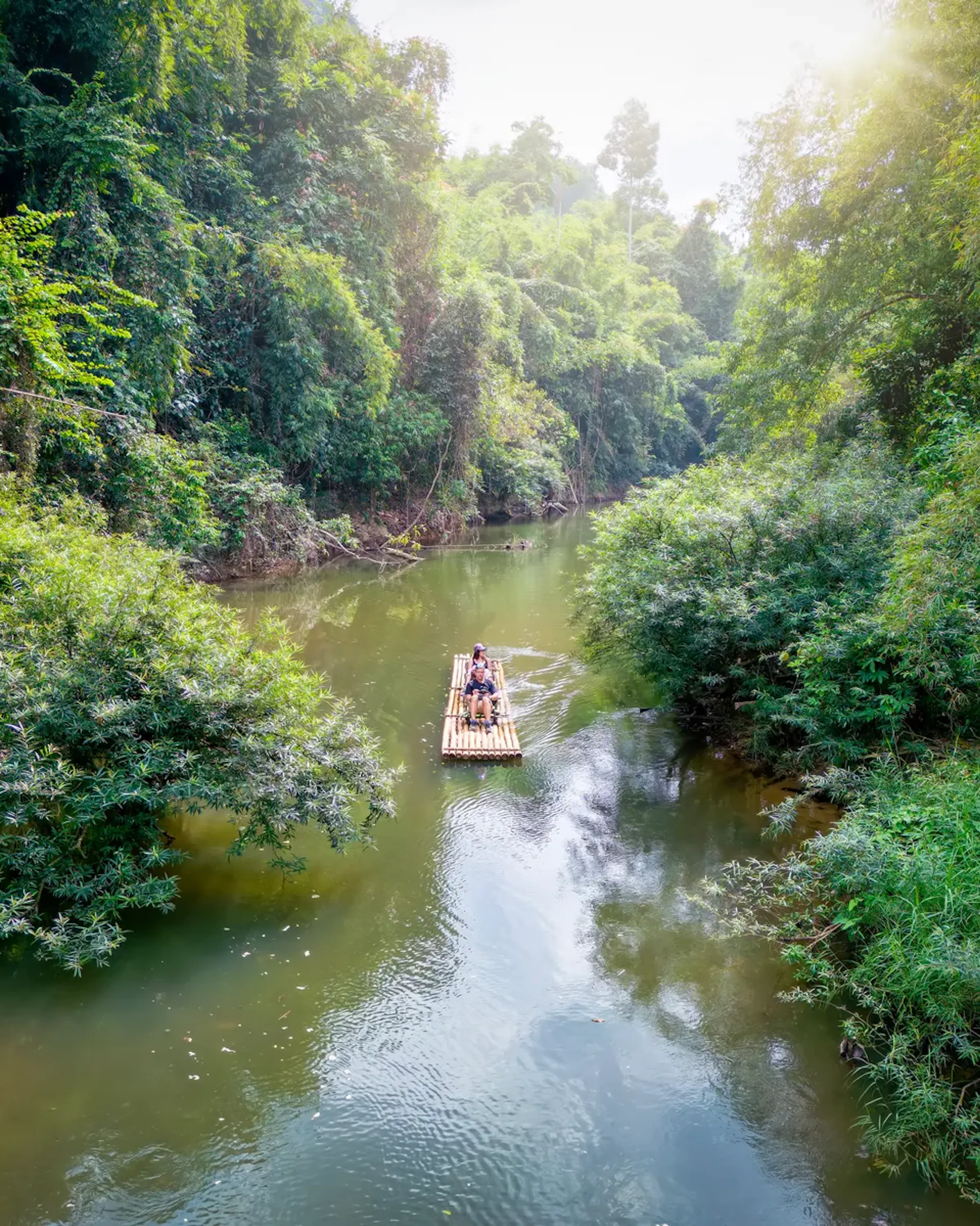 A small bamboo raft carrying two people floats along a calm river surrounded by dense, lush jungle vegetation, with sunlight filtering through the treetops.