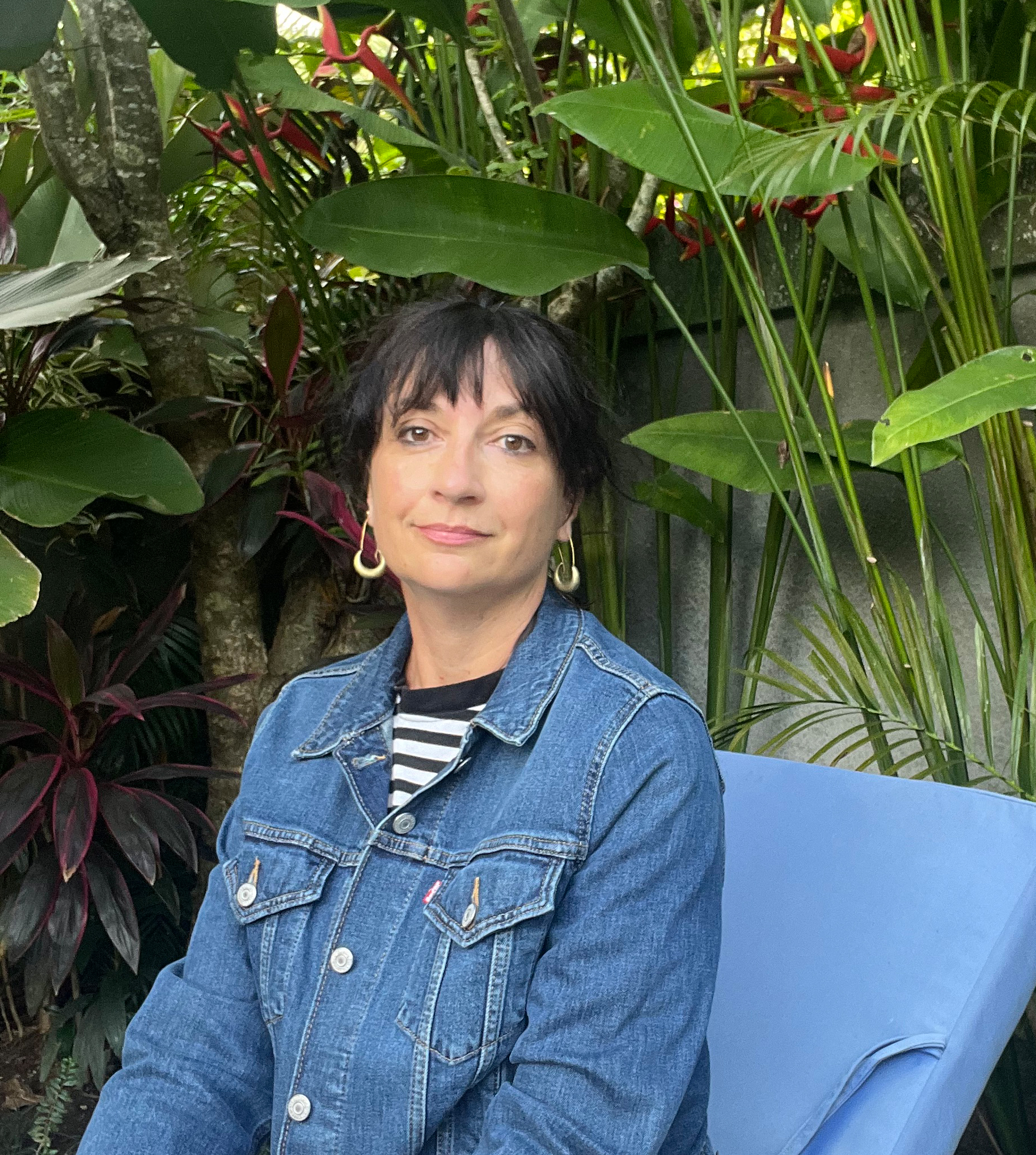 A woman with dark hair and bangs sits on a blue chair in a lush garden filled with tropical plants and red flowers. She is wearing a denim jacket over a black-and-white striped shirt, looking calmly toward the camera.
