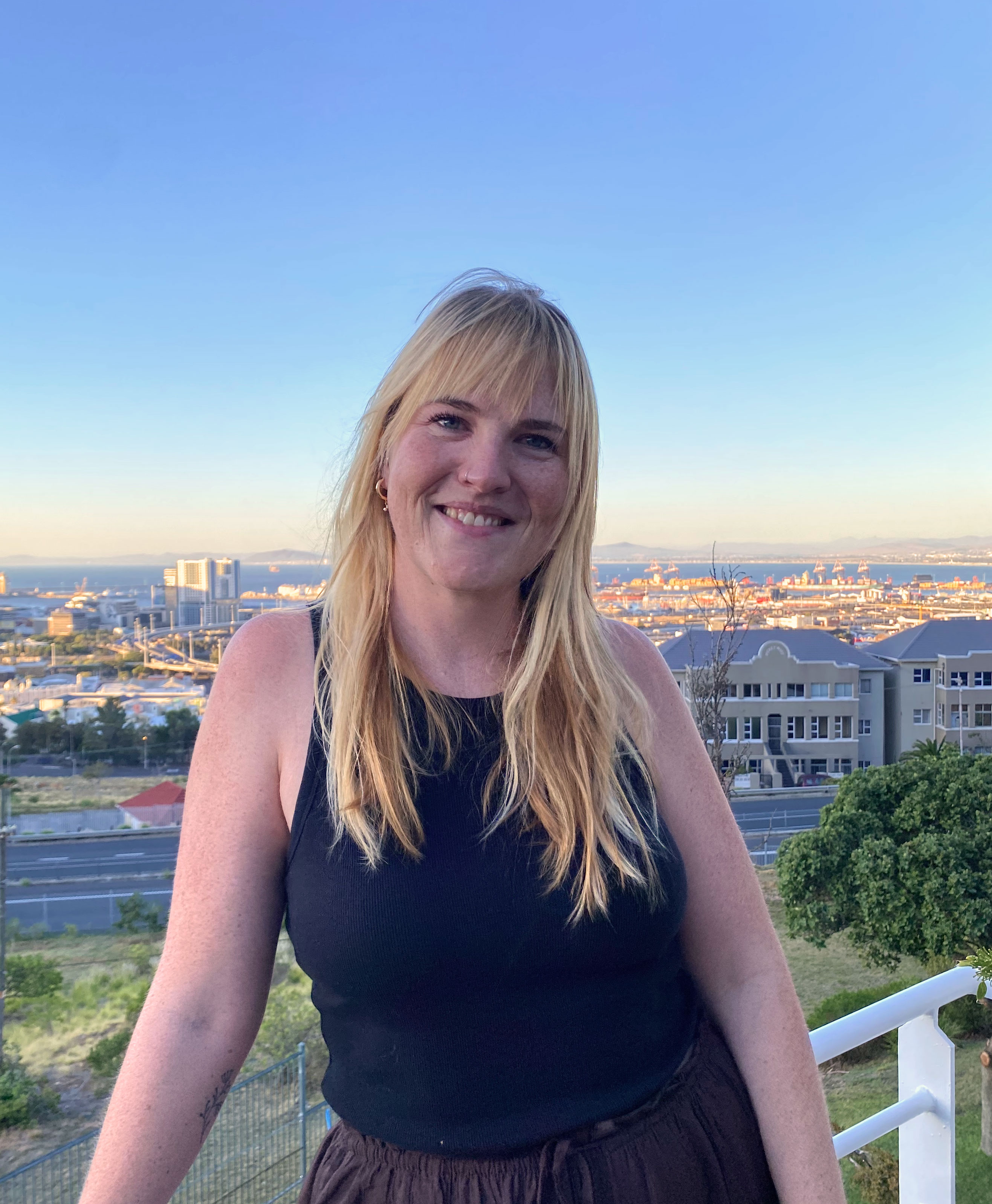 A smiling blonde woman wearing a black sleeveless top stands outdoors on a balcony overlooking a coastal city and harbor at sunset, with buildings, cranes, and mountains visible in the distance under a clear sky.