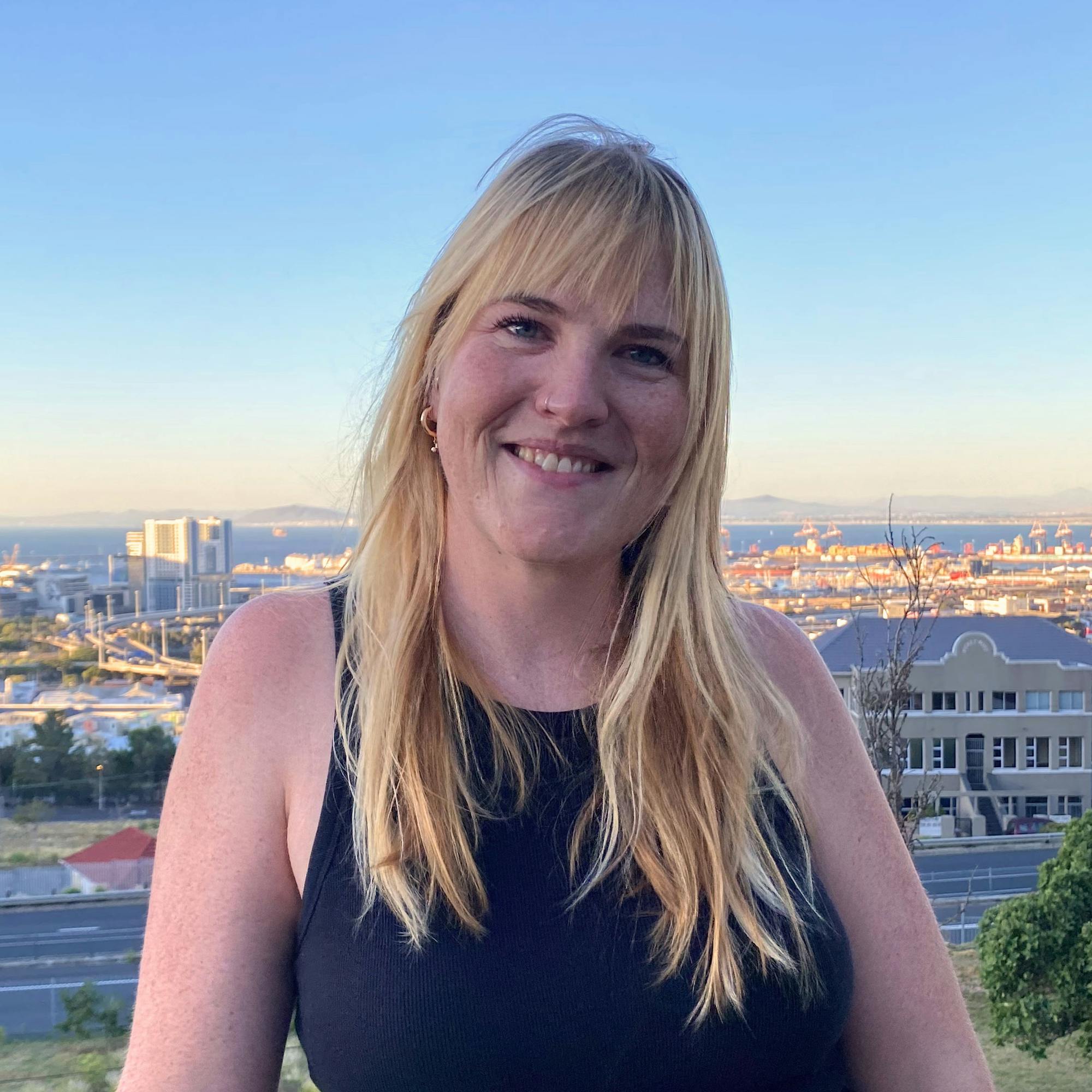 A smiling blonde woman wearing a black sleeveless top stands outdoors on a balcony overlooking a coastal city and harbor at sunset, with buildings, cranes, and mountains visible in the distance under a clear sky.