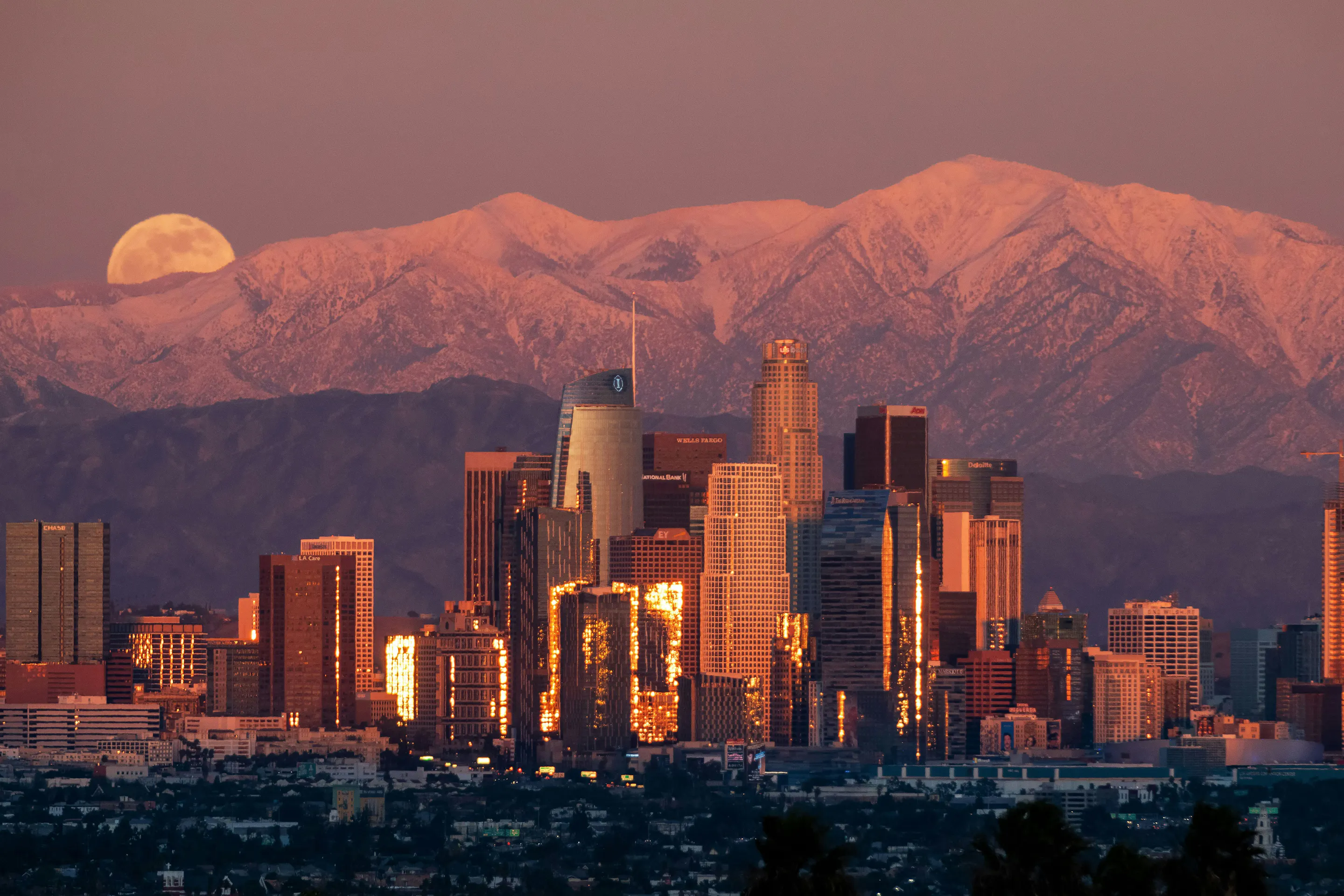 Santiago’s towers glow at dusk beneath a rising moon, with the Andes peaks tinted pink behind them softly.