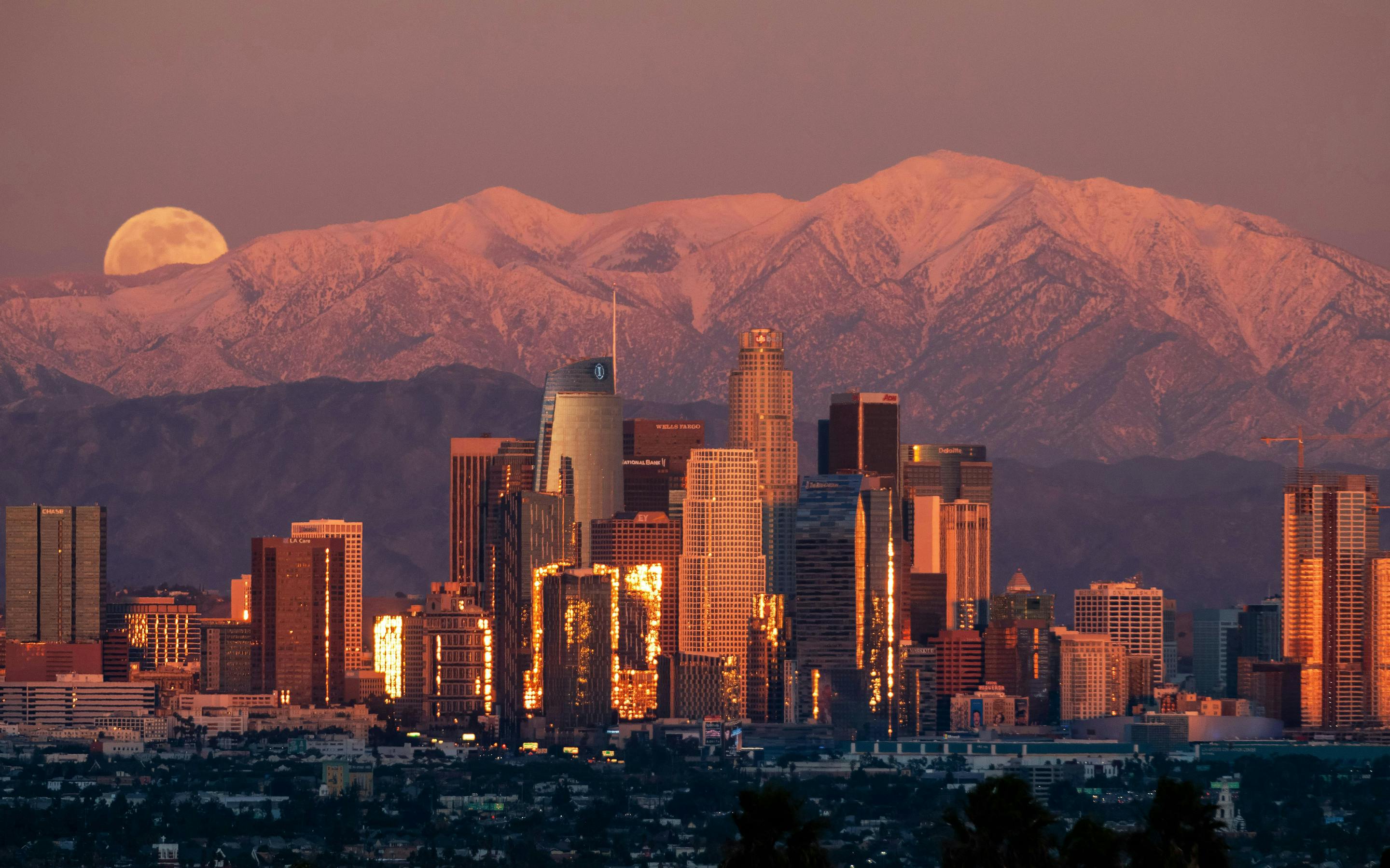 Santiago’s towers glow at dusk beneath a rising moon, with the Andes peaks tinted pink behind them softly.
