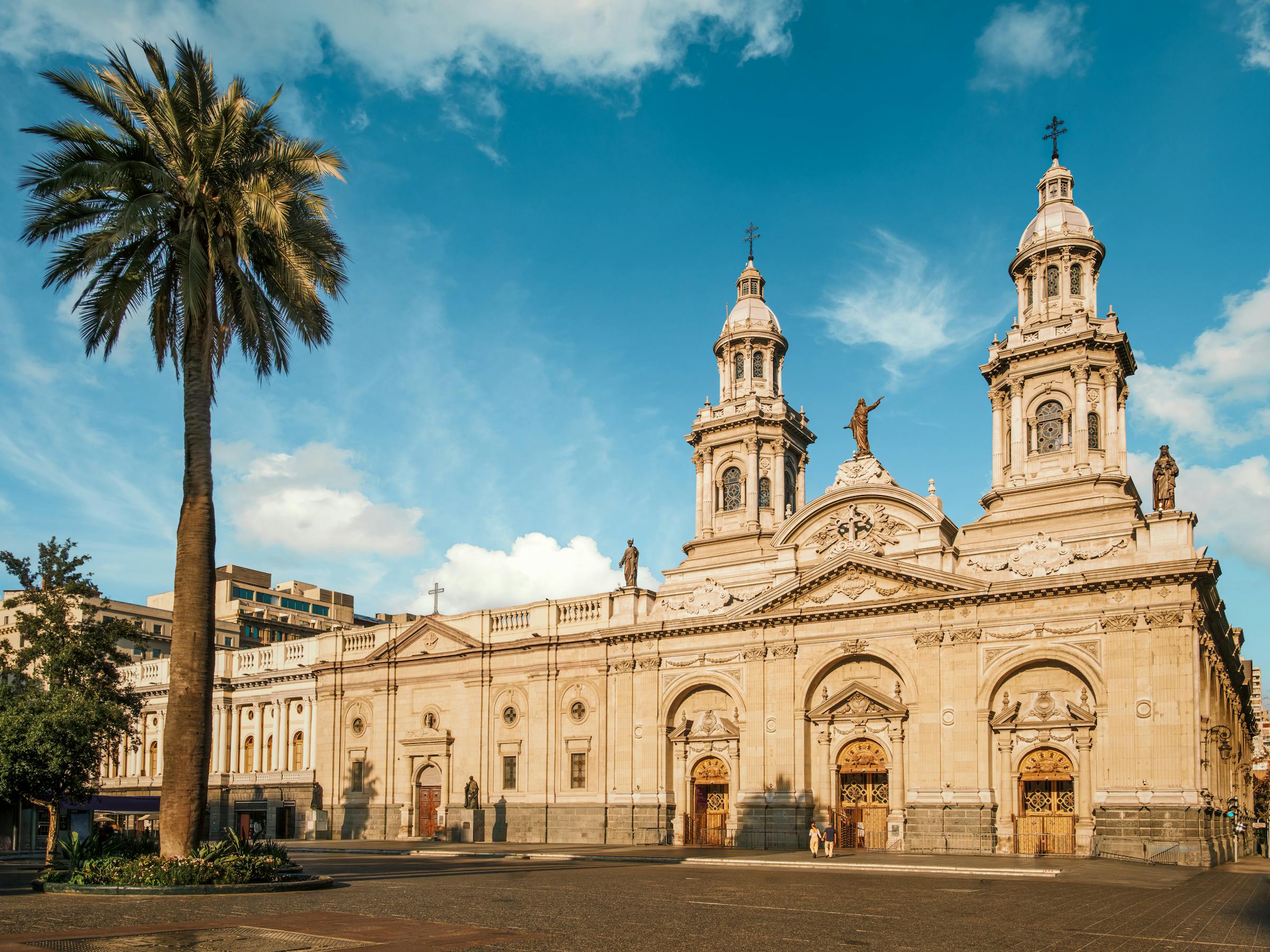 The Metropolitan Cathedral of Santiago stands in Plaza de Armas, with a palm framing its twin towers in sun.