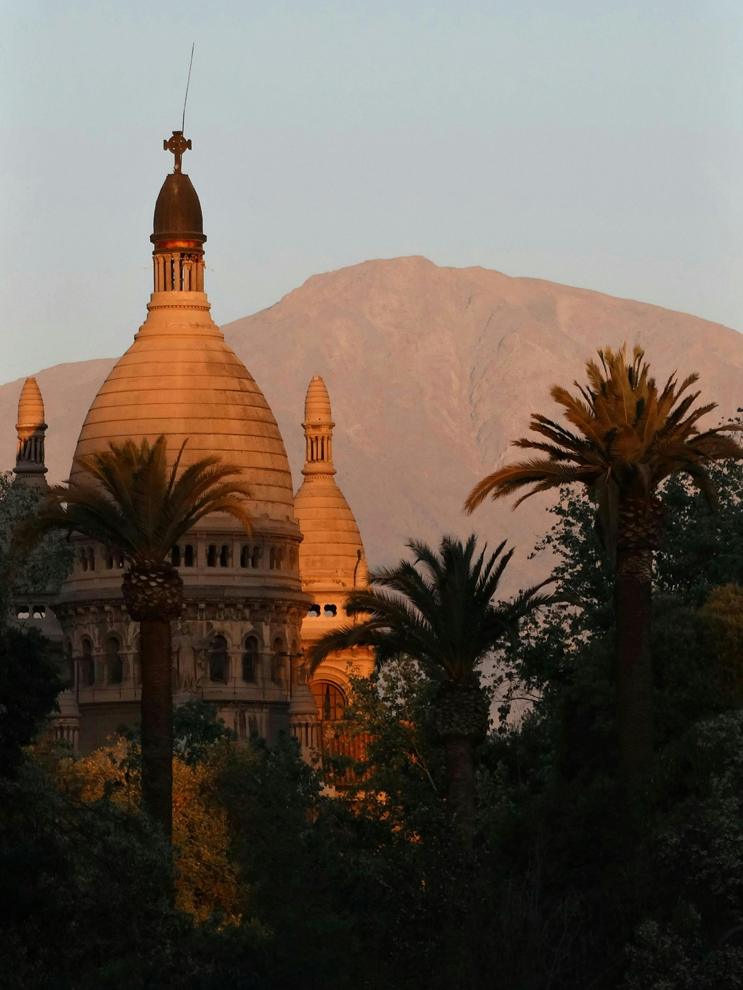 A domed historic building rises among palm trees in Santiago at sunset, with the Andes in soft haze beyond.