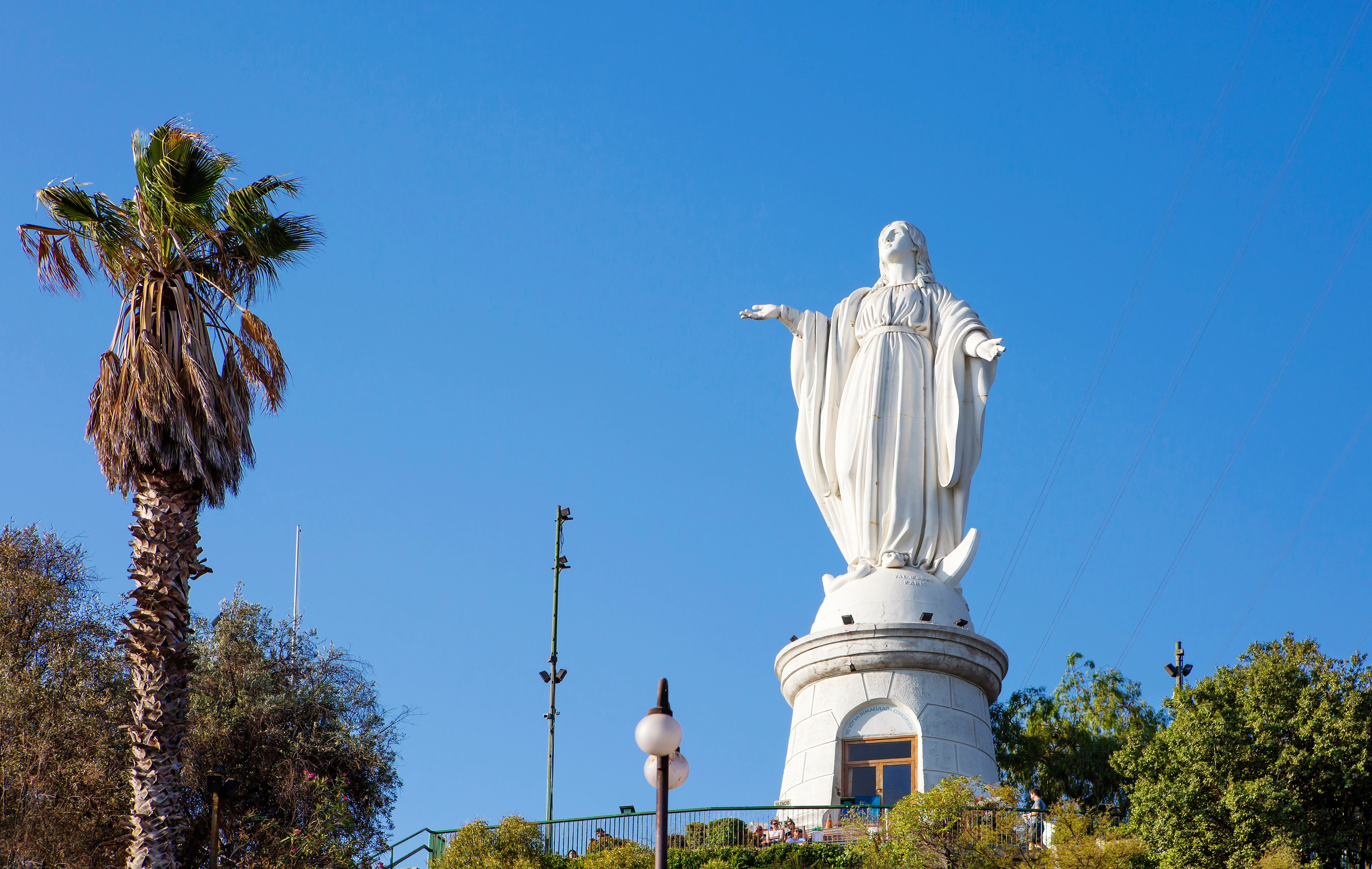 The white statue of the Virgin Mary rises above Cerro San Cristóbal, with palms under a clear blue sky above.