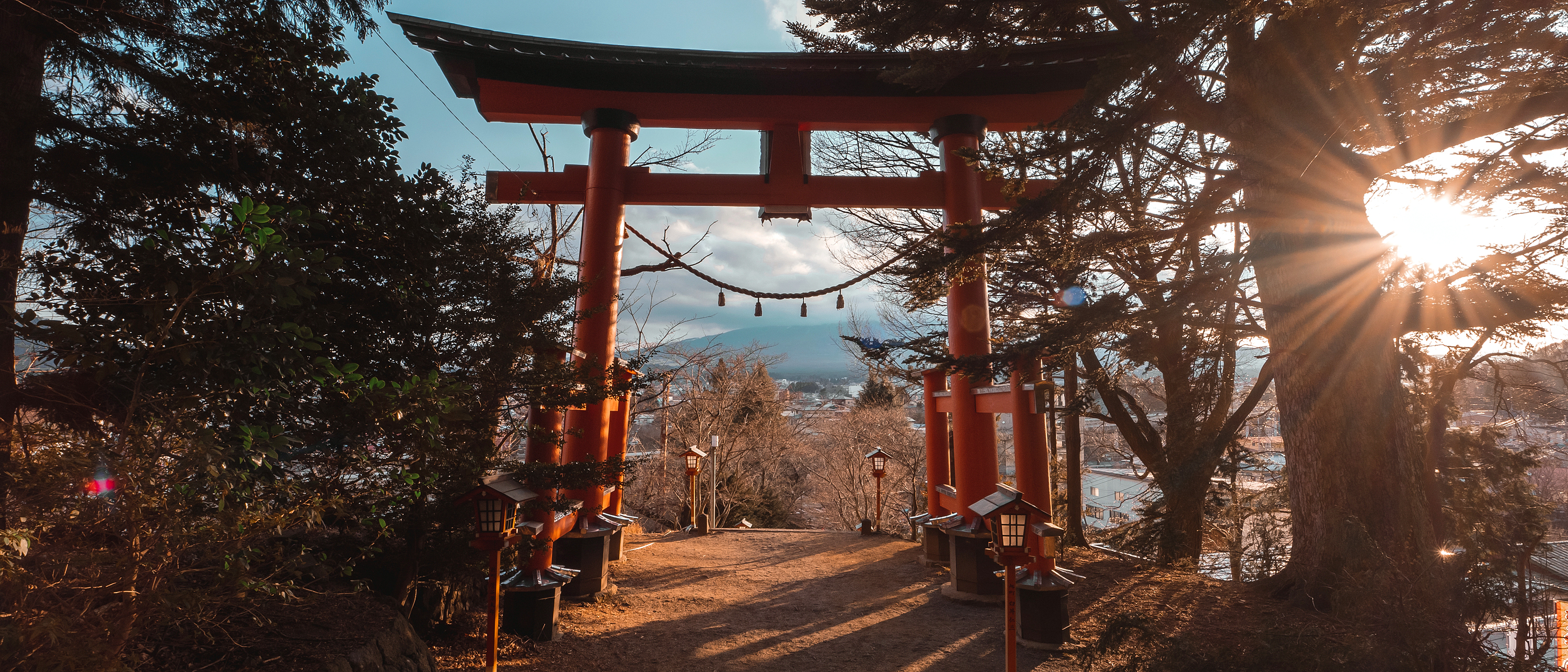 A red torii gate stands near Lake Kawaguchiko, with sunrise light flaring through trees along the path below.
