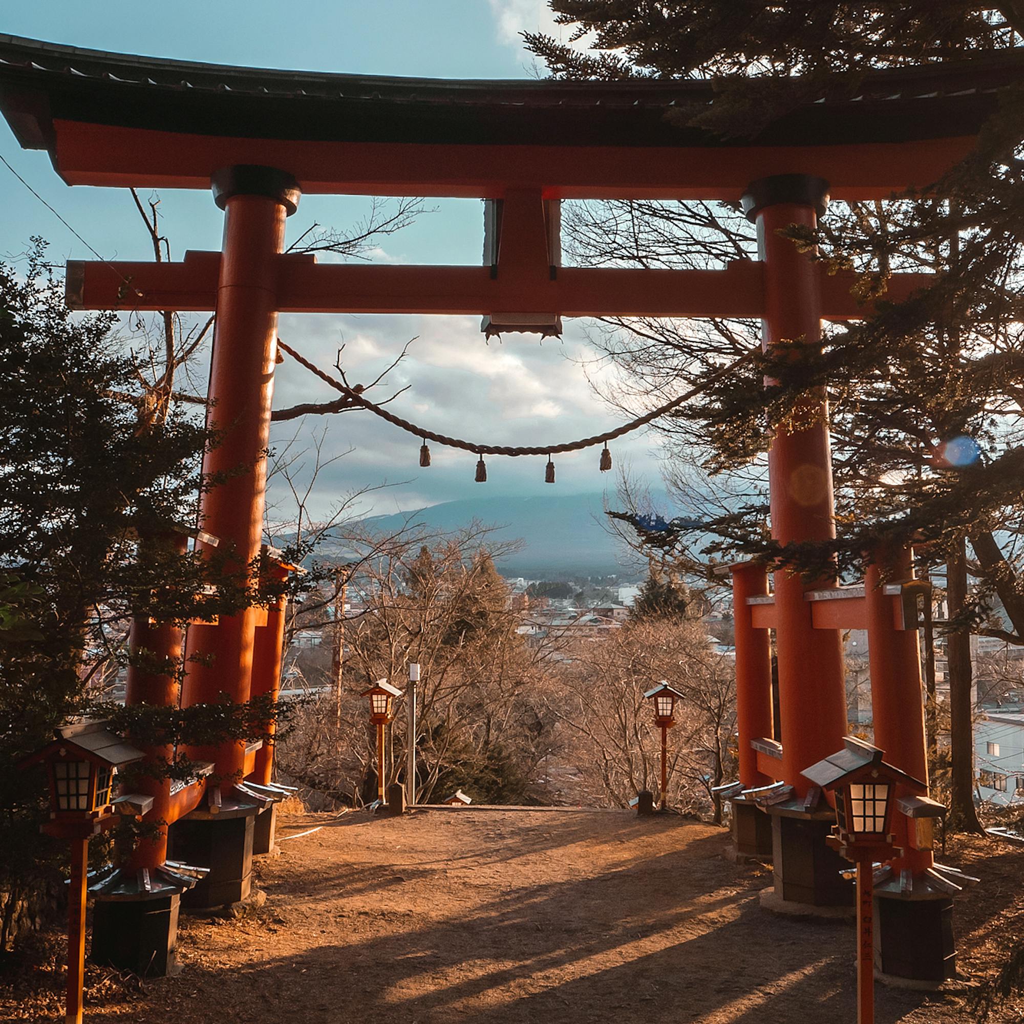 A red torii gate stands near Lake Kawaguchiko, with sunrise light flaring through trees along the path below.