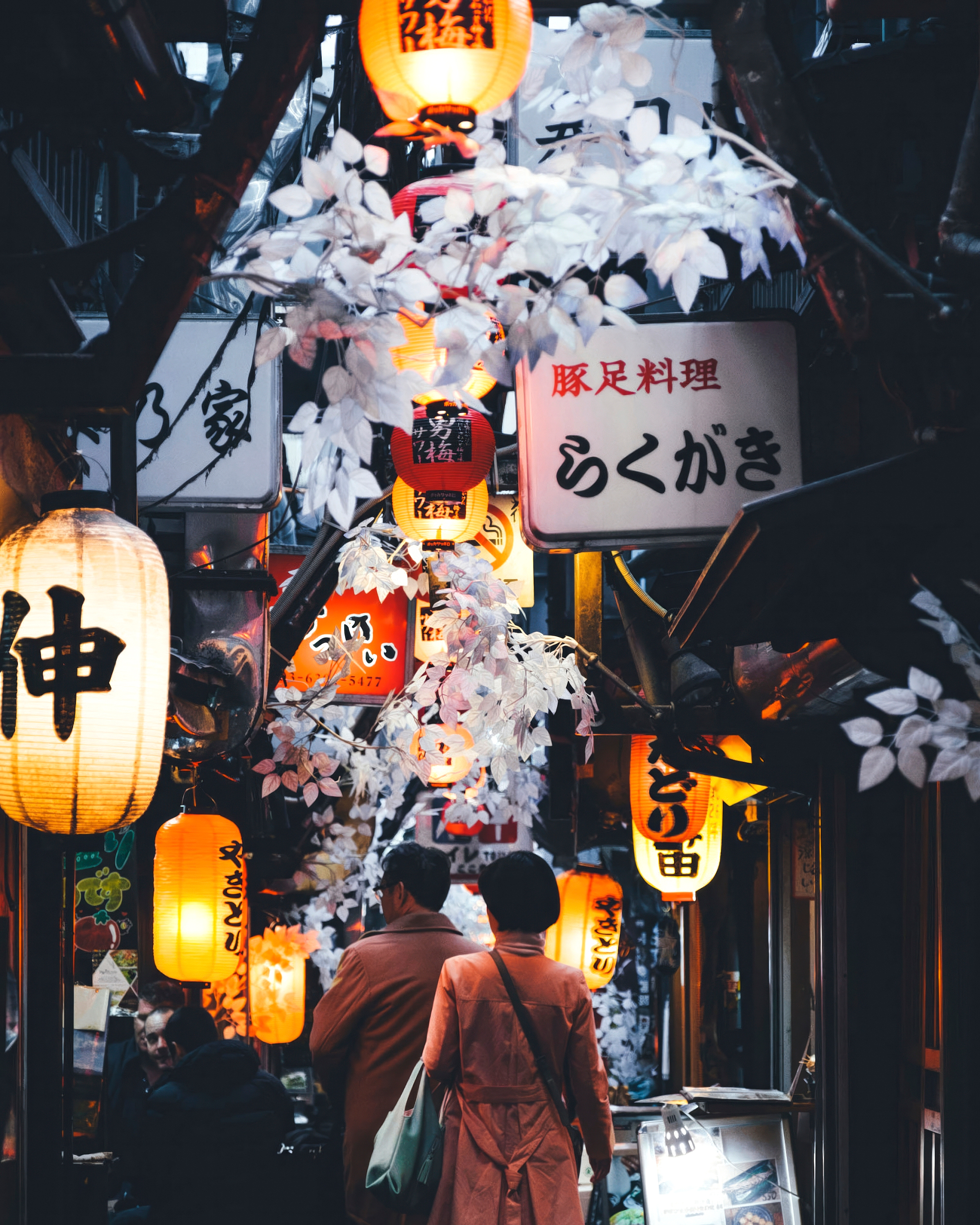 A narrow Tokyo alley glows with lanterns and paper signs at night, with diners walking between small bars.