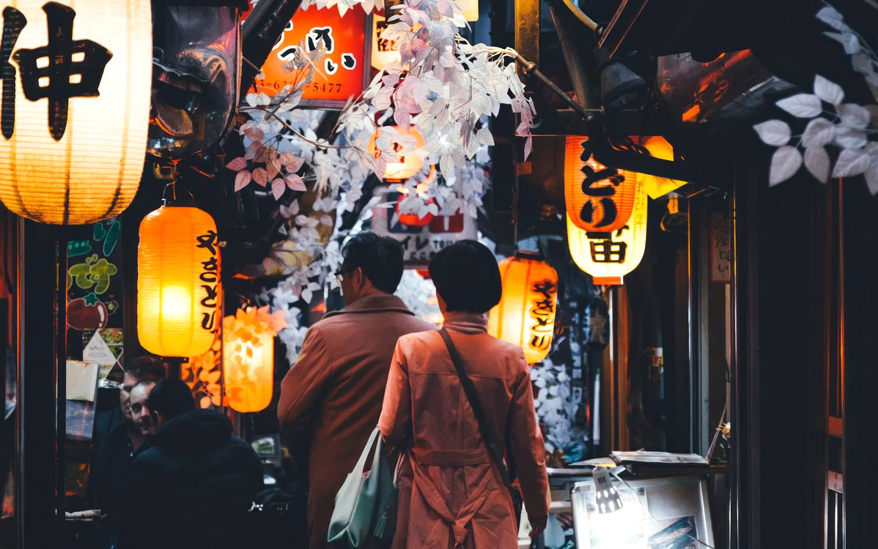A narrow Tokyo alley glows with lanterns and paper signs at night, with diners walking between small bars.