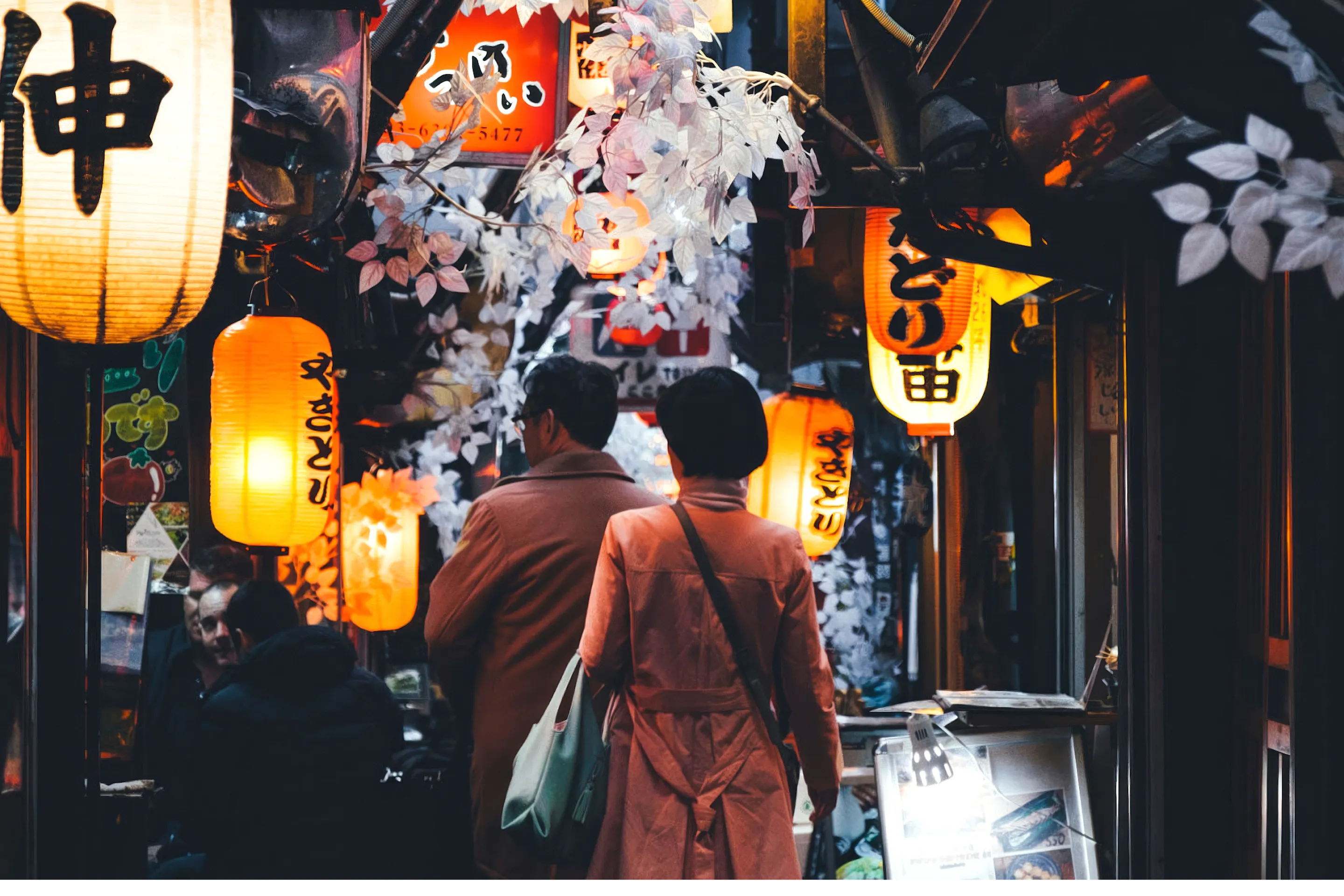 A narrow Tokyo alley glows with lanterns and paper signs at night, with diners walking between small bars.