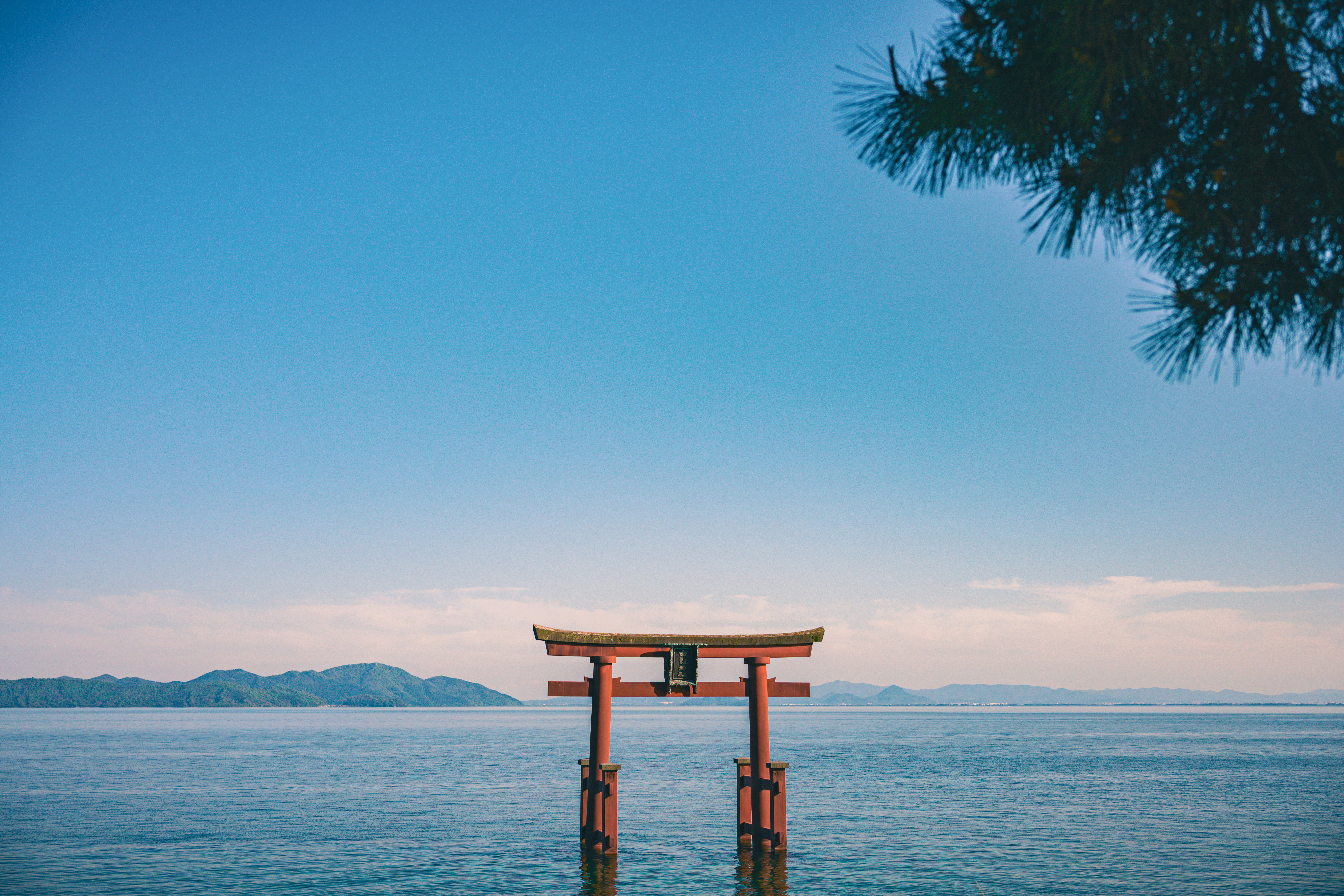 A lone torii gate stands in shallow water under a clear blue sky, with distant islands low on the horizon.