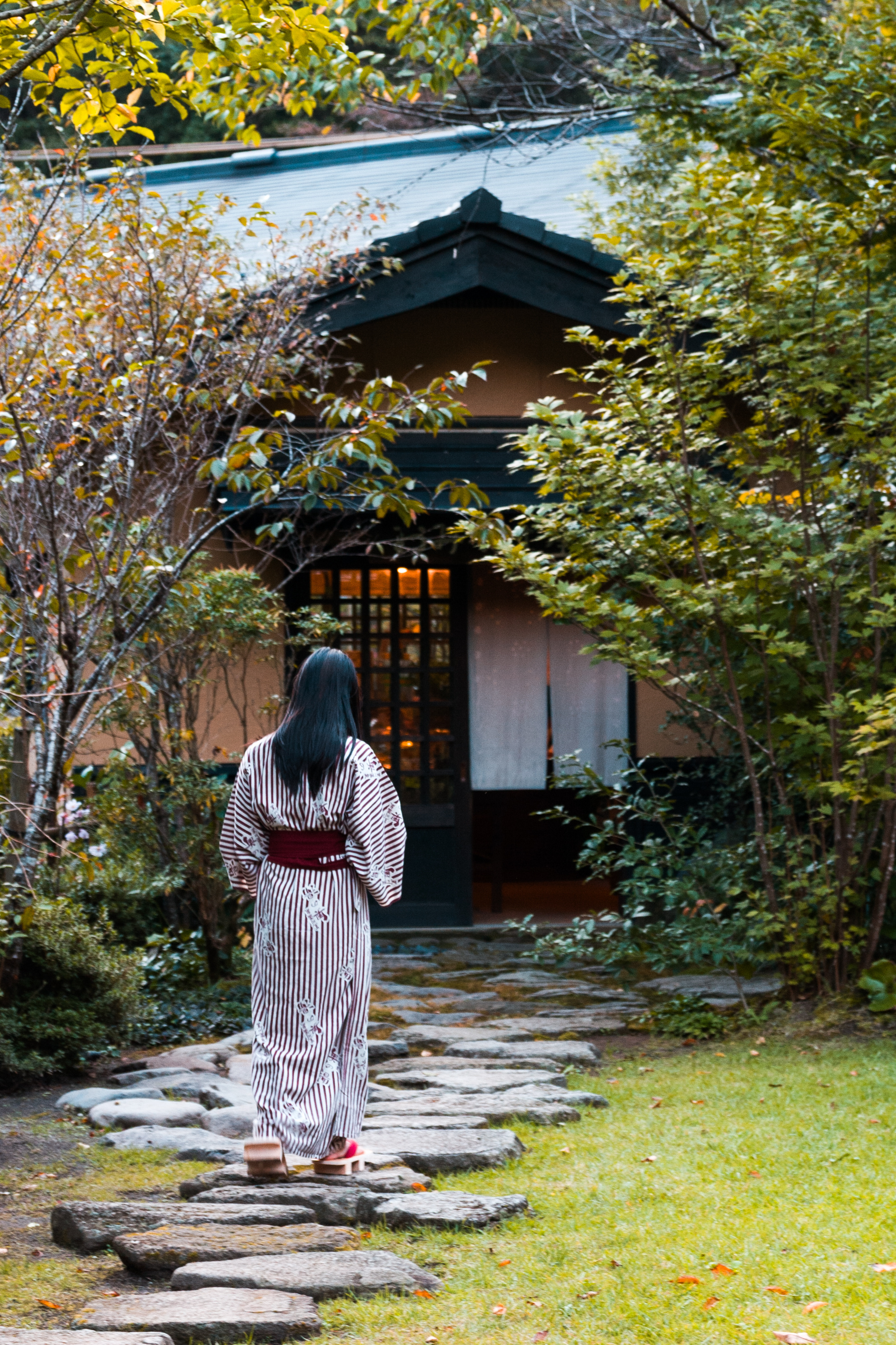A woman in a striped yukata walks on stepping stones at Kurokawa Onsen, surrounded by trees and a wooden inn.