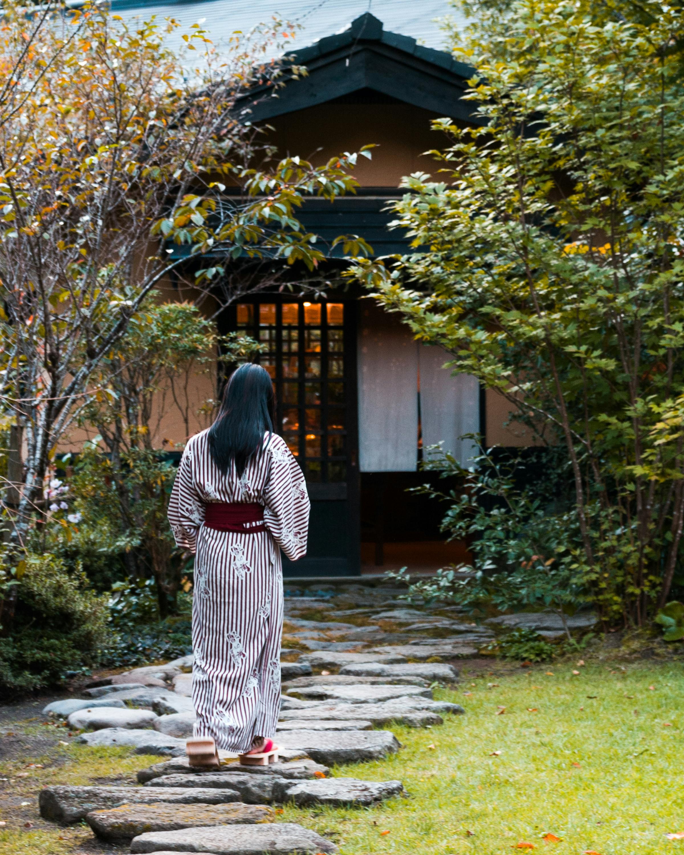 A woman in a striped yukata walks on stepping stones at Kurokawa Onsen, surrounded by trees and a wooden inn.