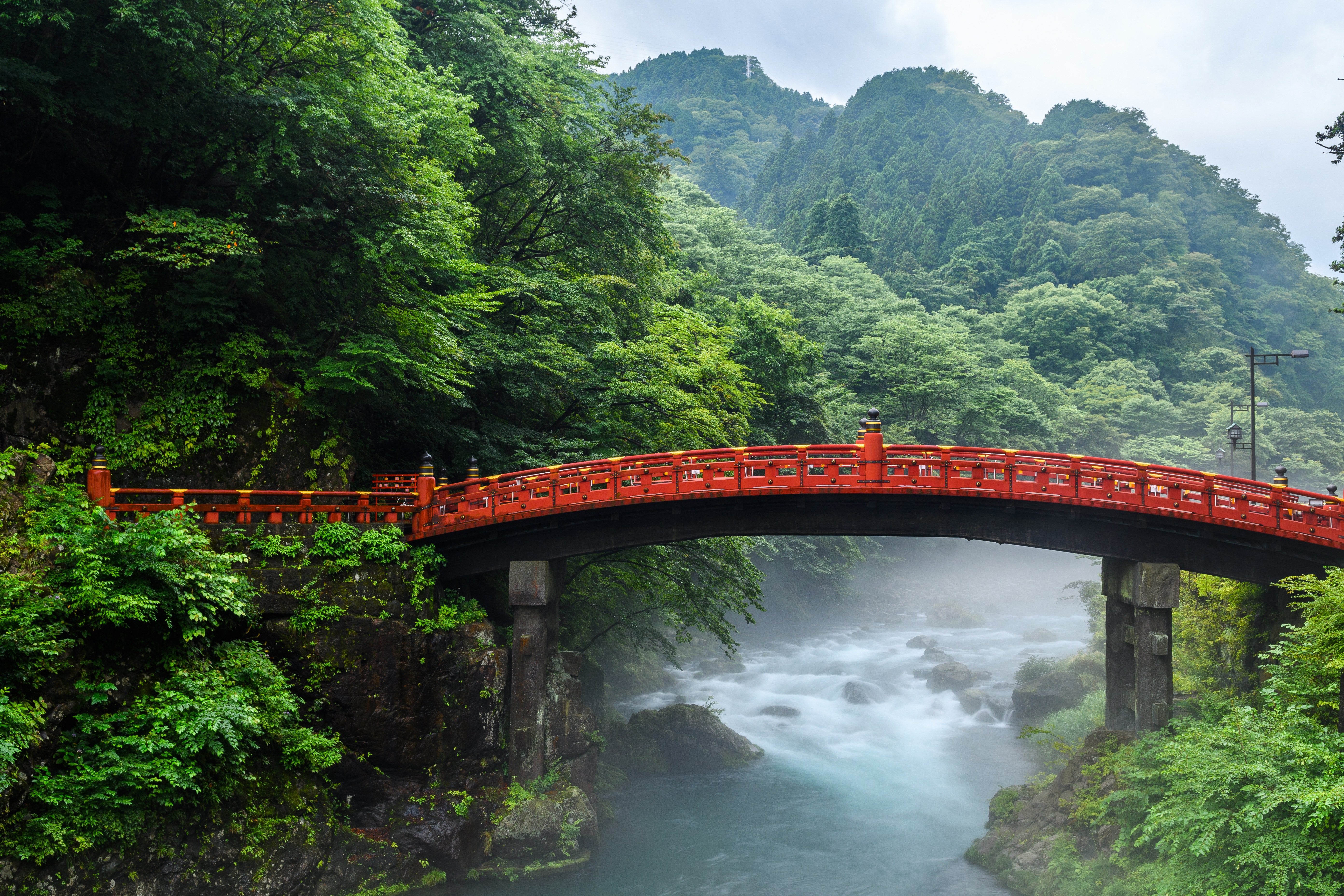 The red Shinkyo Bridge in Nikko spans a misty river gorge in forest, with rushing water below and hills beyond.