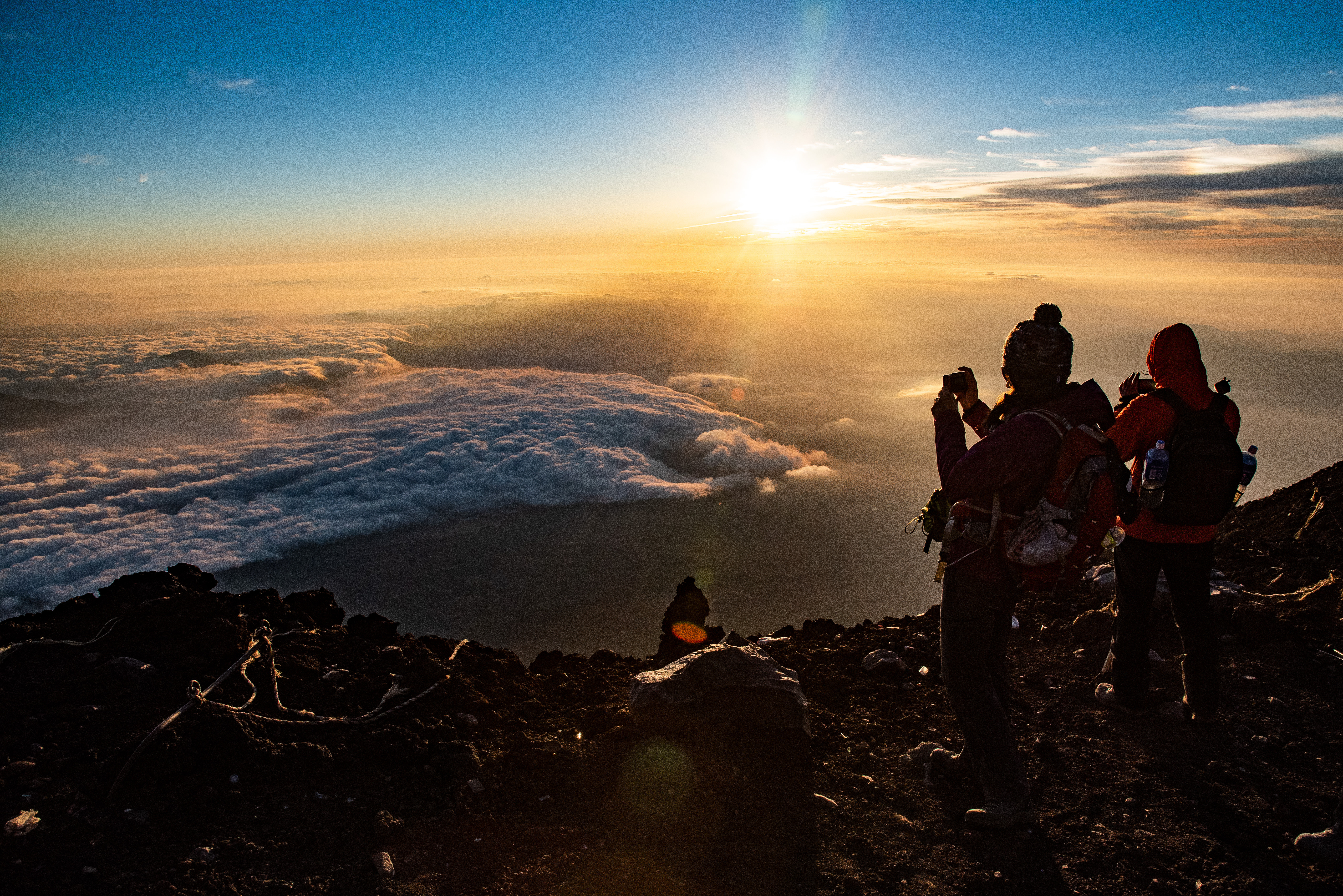 Hikers watch sunrise near Mount Fuji from a rocky peak above a sea of clouds, with golden light on the horizon.