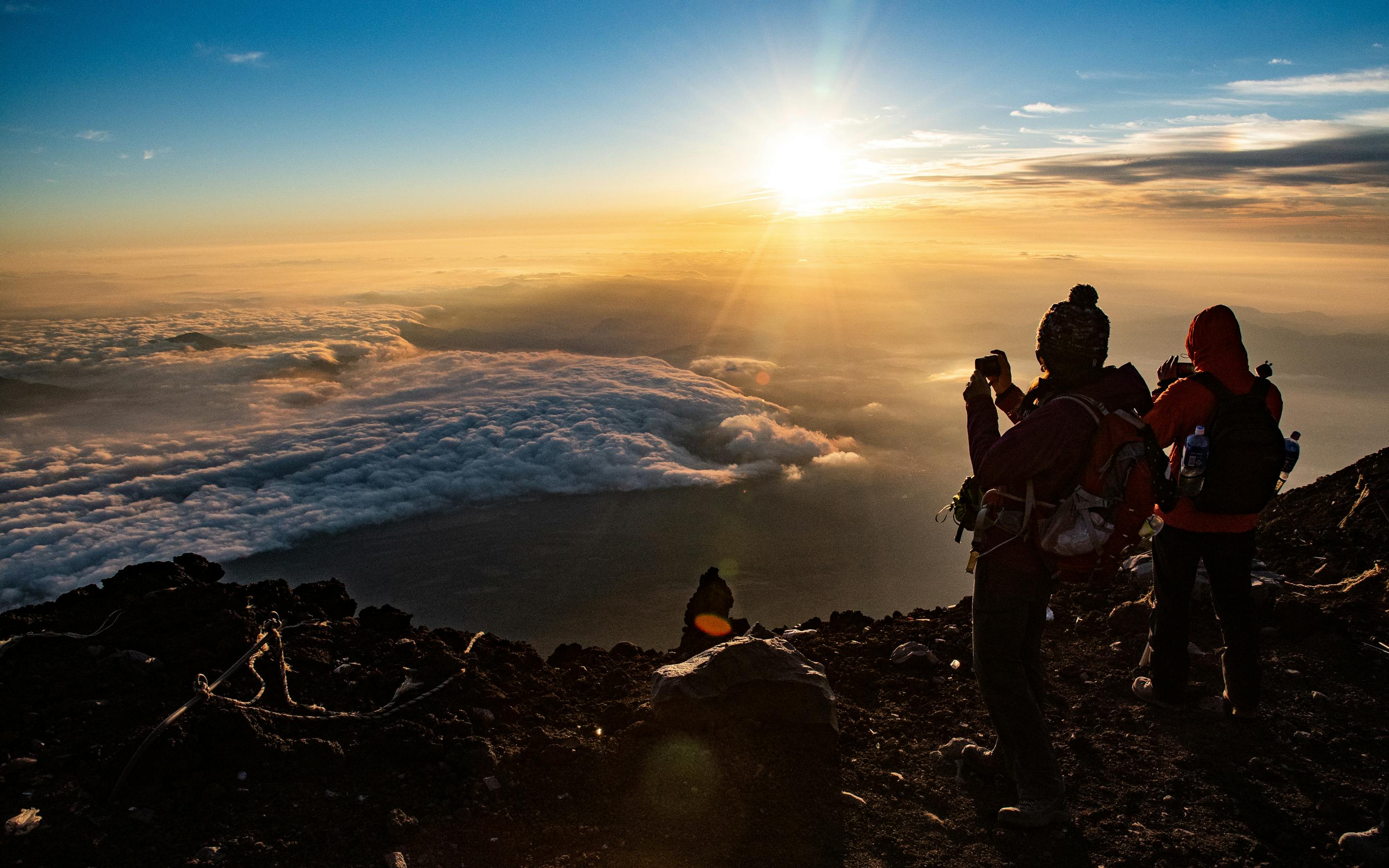 Hikers watch sunrise near Mount Fuji from a rocky peak above a sea of clouds, with golden light on the horizon.