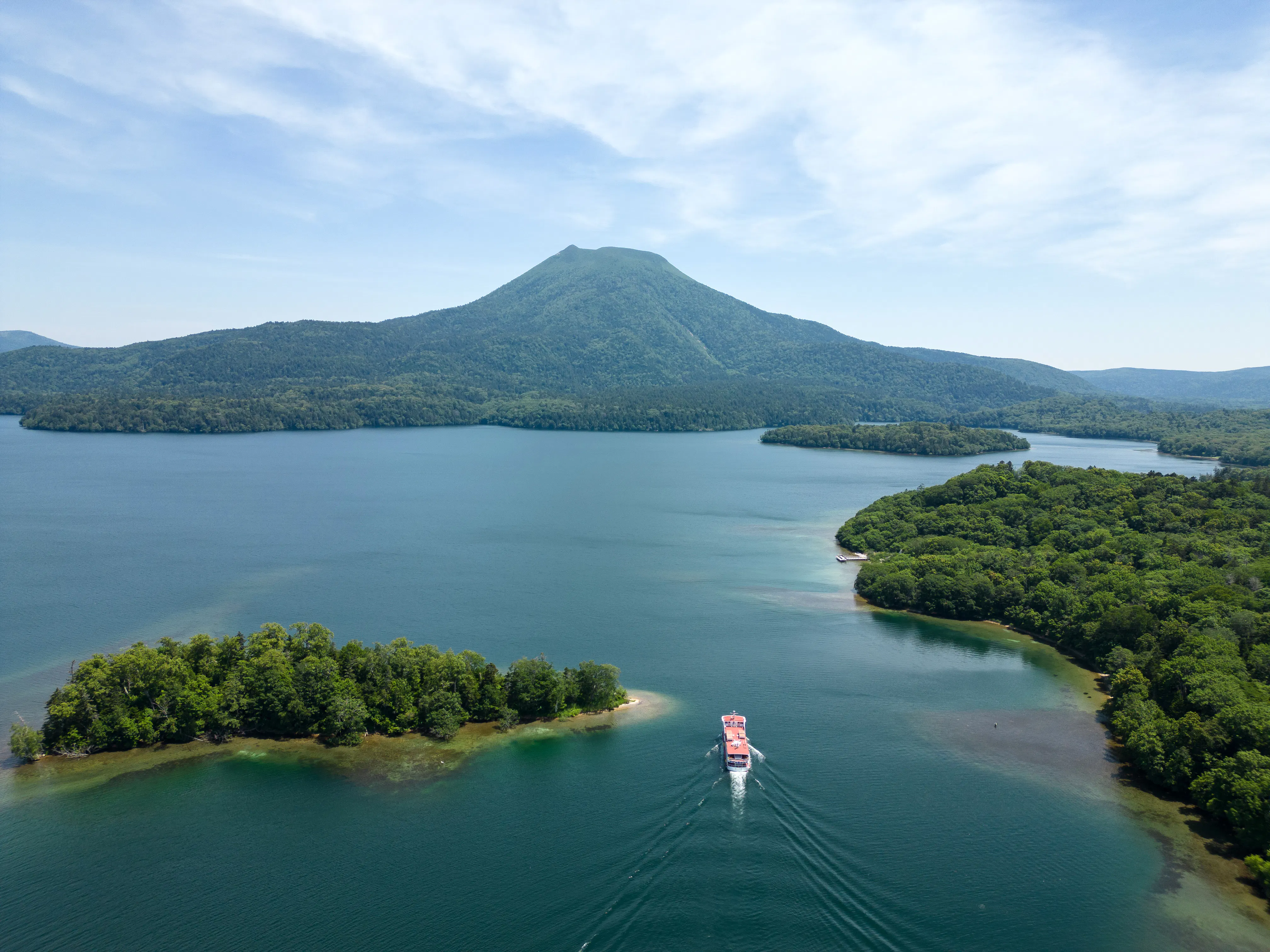 A tour boat cruises across Lake Akan in Hokkaido, with Mount Oakan volcano rising above forested shores behind.