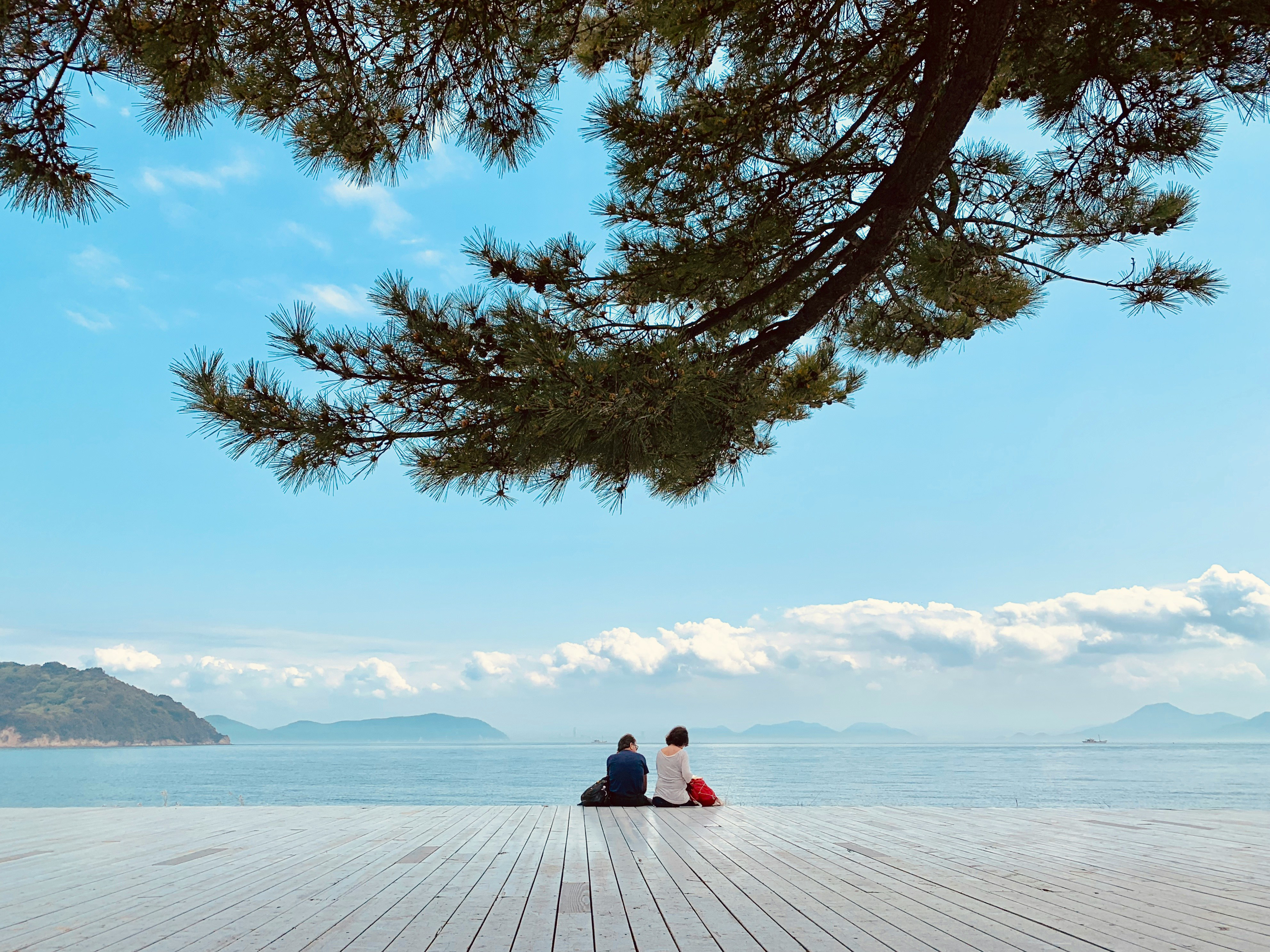 Two people sit on a deck on Naoshima Island in Kagawa Prefecture, Shikoku, looking out over calm blue water.