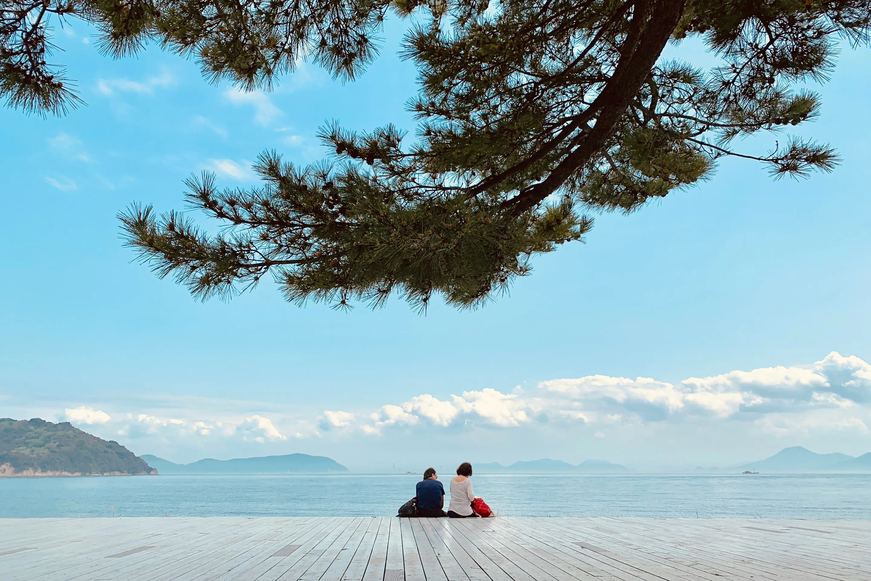 Two people sit on a deck on Naoshima Island in Kagawa Prefecture, Shikoku, looking out over calm blue water.
