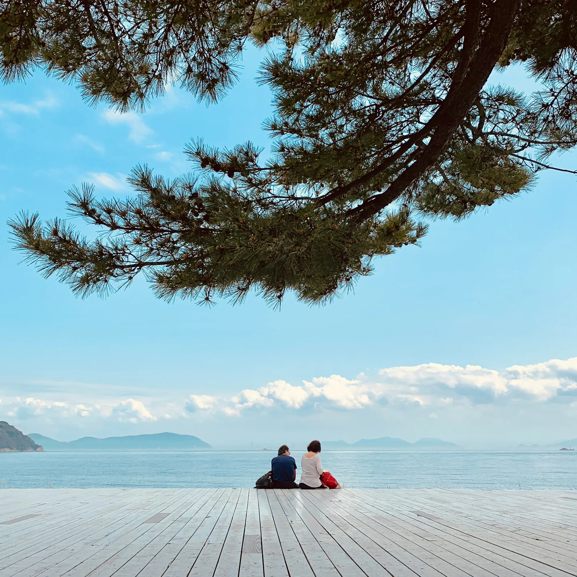 Two people sit on a deck on Naoshima Island in Kagawa Prefecture, Shikoku, looking out over calm blue water.