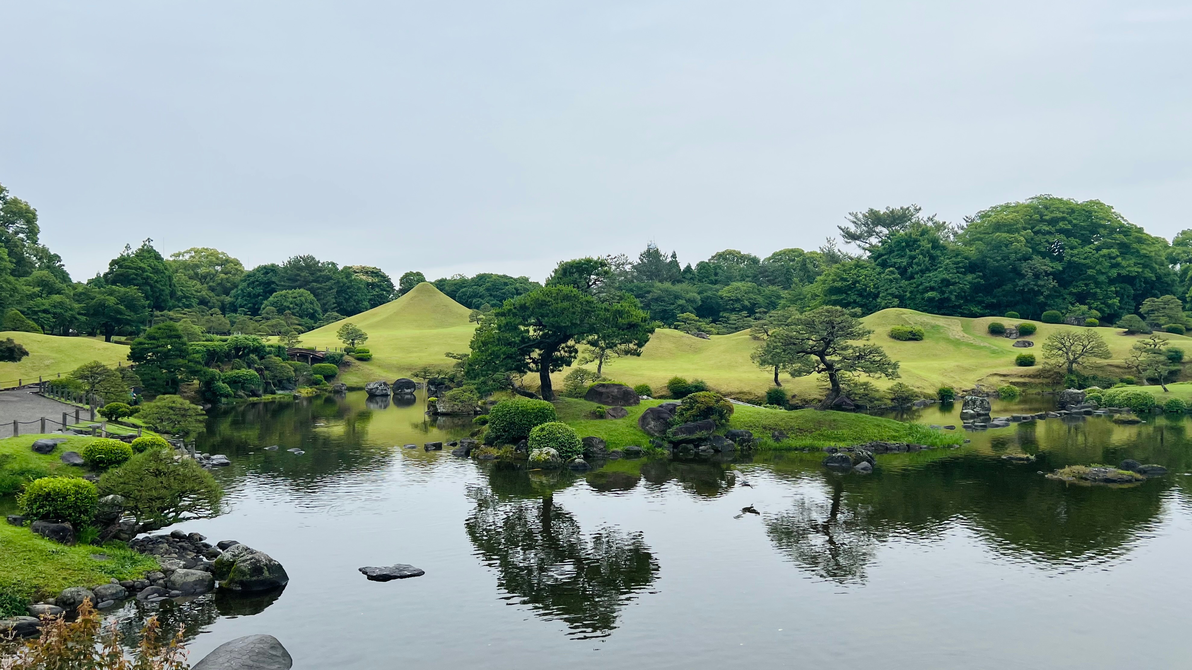 A manicured Japanese garden surrounds a still pond, with clipped green hills and trees reflected in the water.