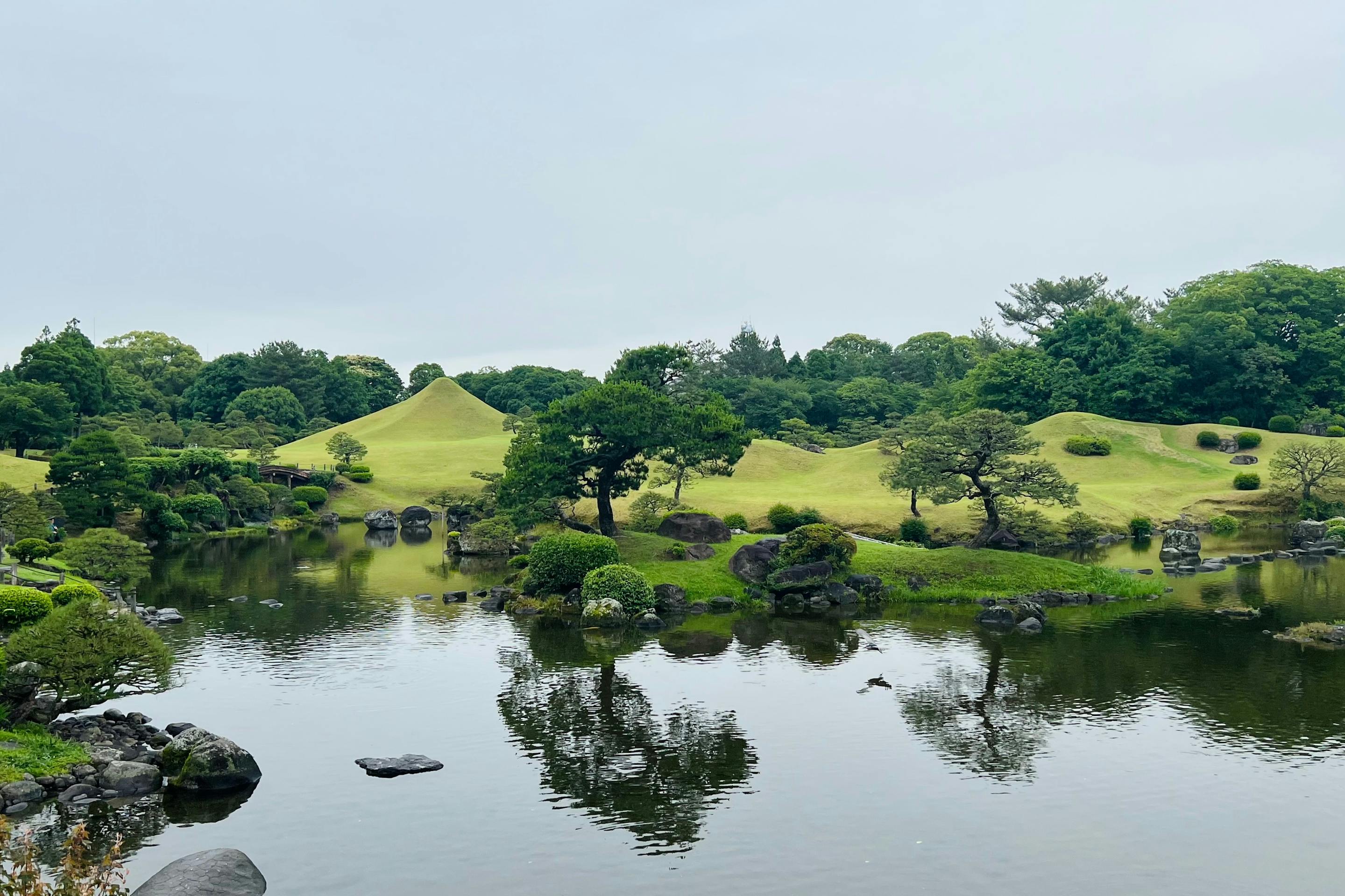 A manicured Japanese garden surrounds a still pond, with clipped green hills and trees reflected in the water.
