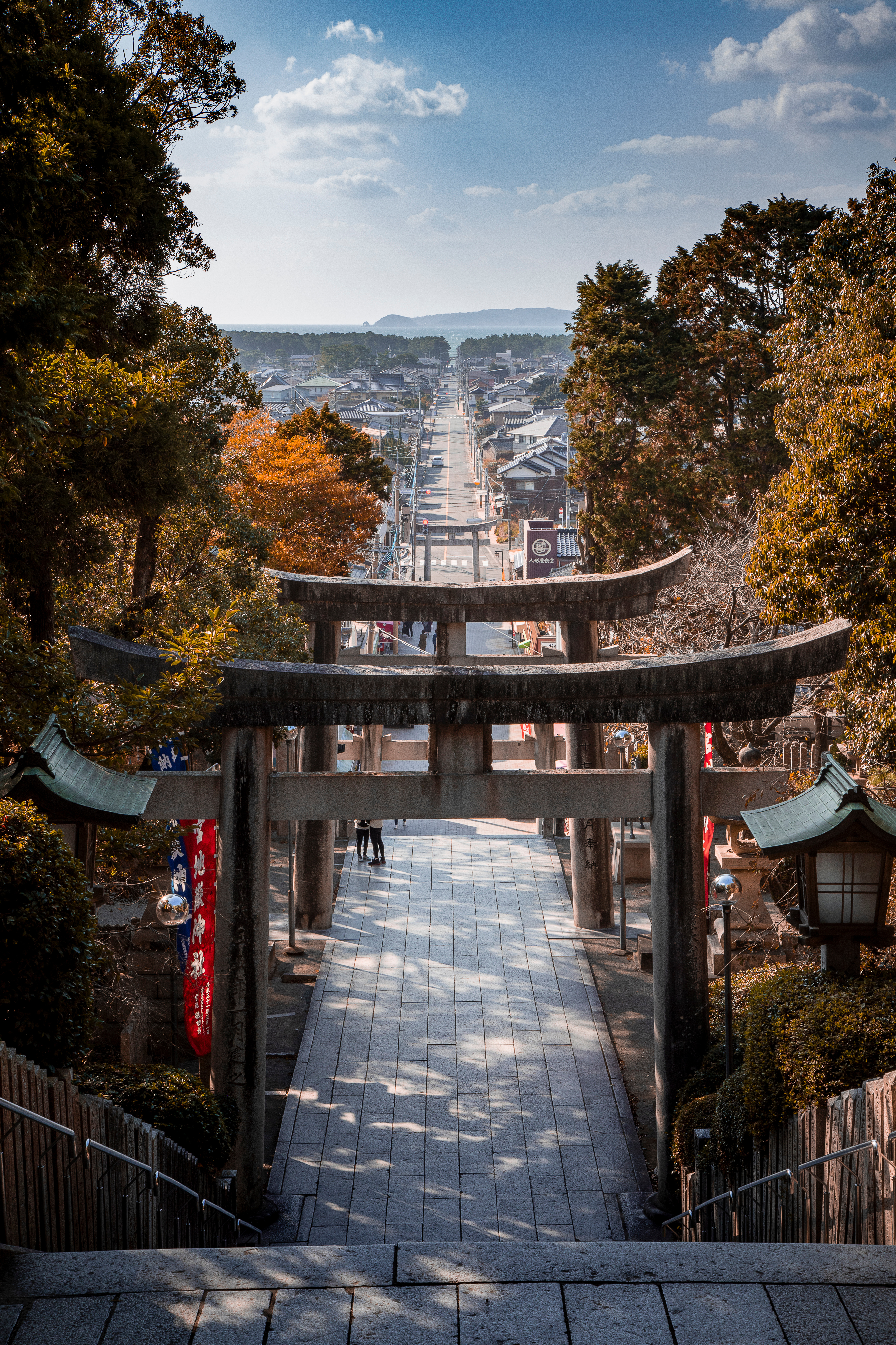 A torii gate frames stone steps descending toward a town, with autumn trees lining the shrine approach below.