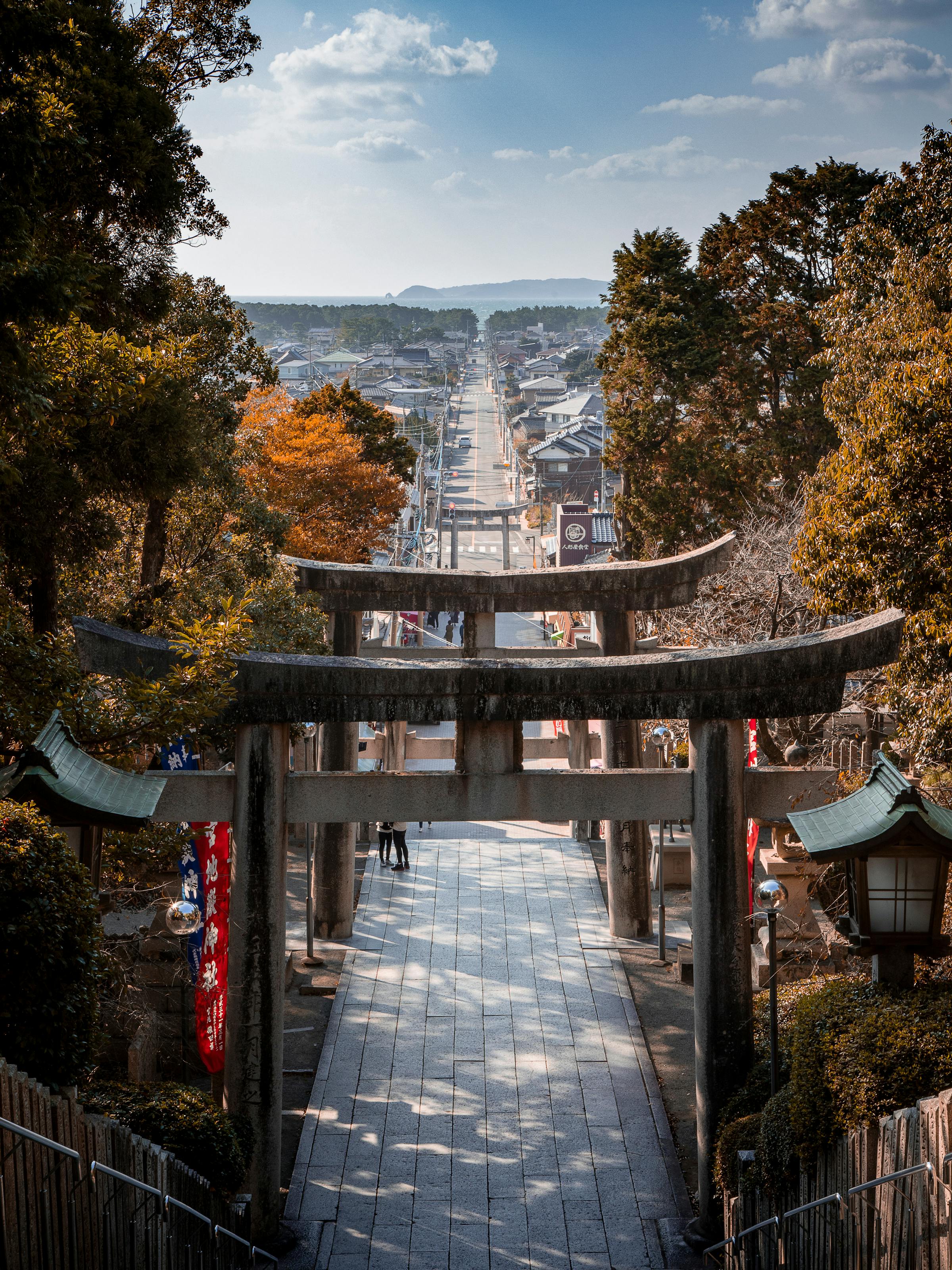 A torii gate frames stone steps descending toward a town, with autumn trees lining the shrine approach below.
