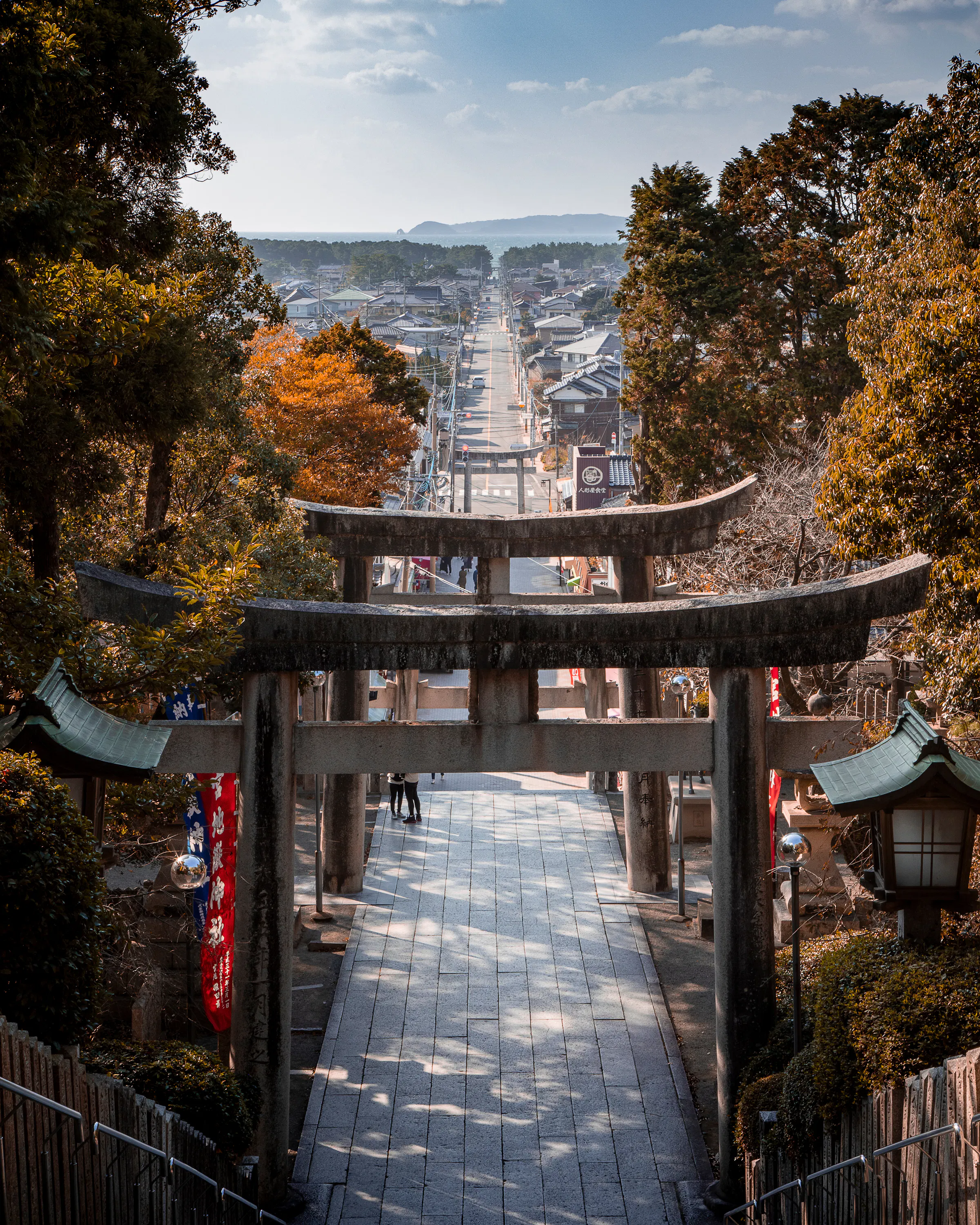 A torii gate frames stone steps descending toward a town, with autumn trees lining the shrine approach below.
