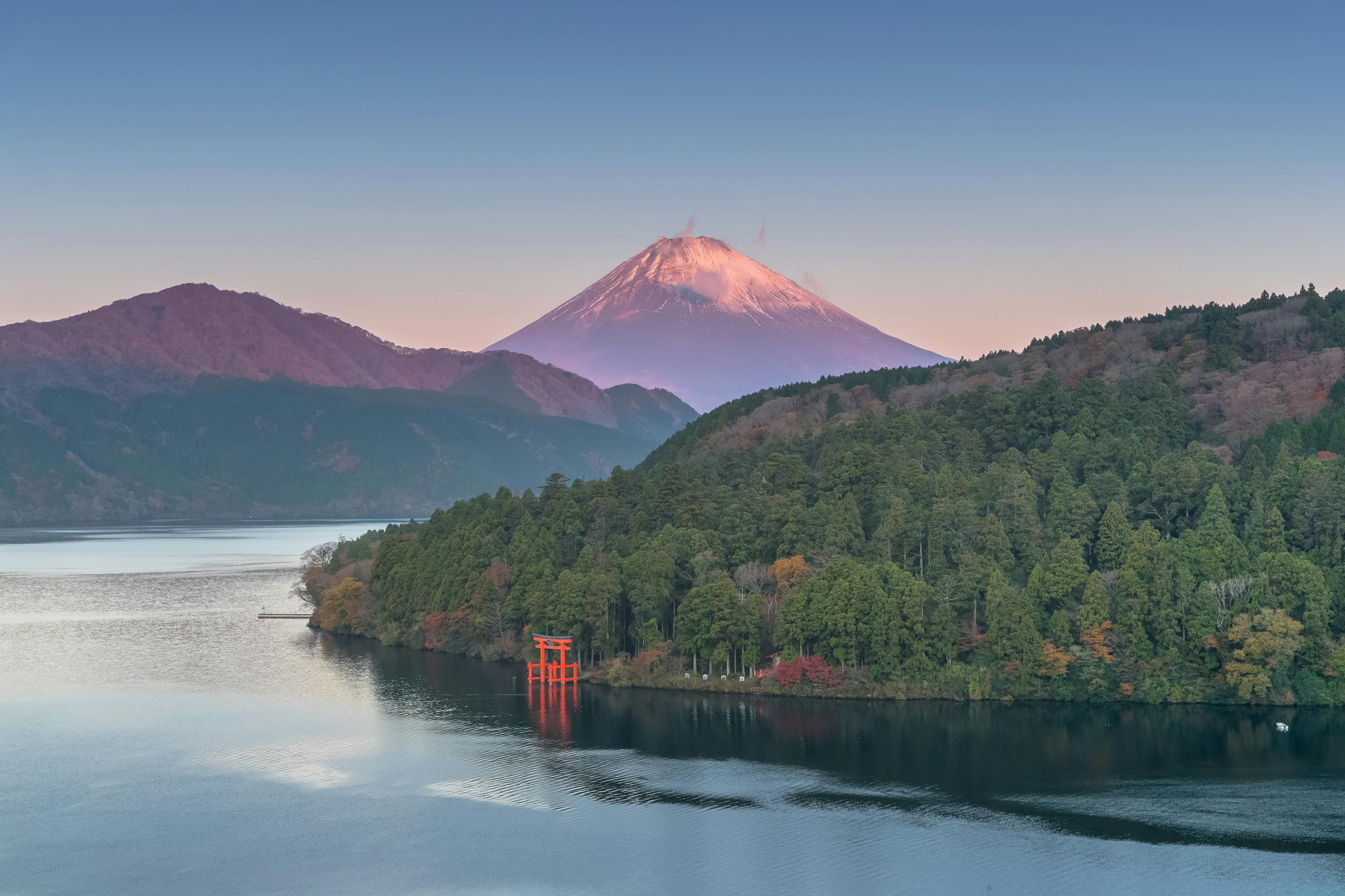 Mount Fuji rises beyond Lake Ashi at sunrise, with a red torii near the shore and forested hills in autumn.