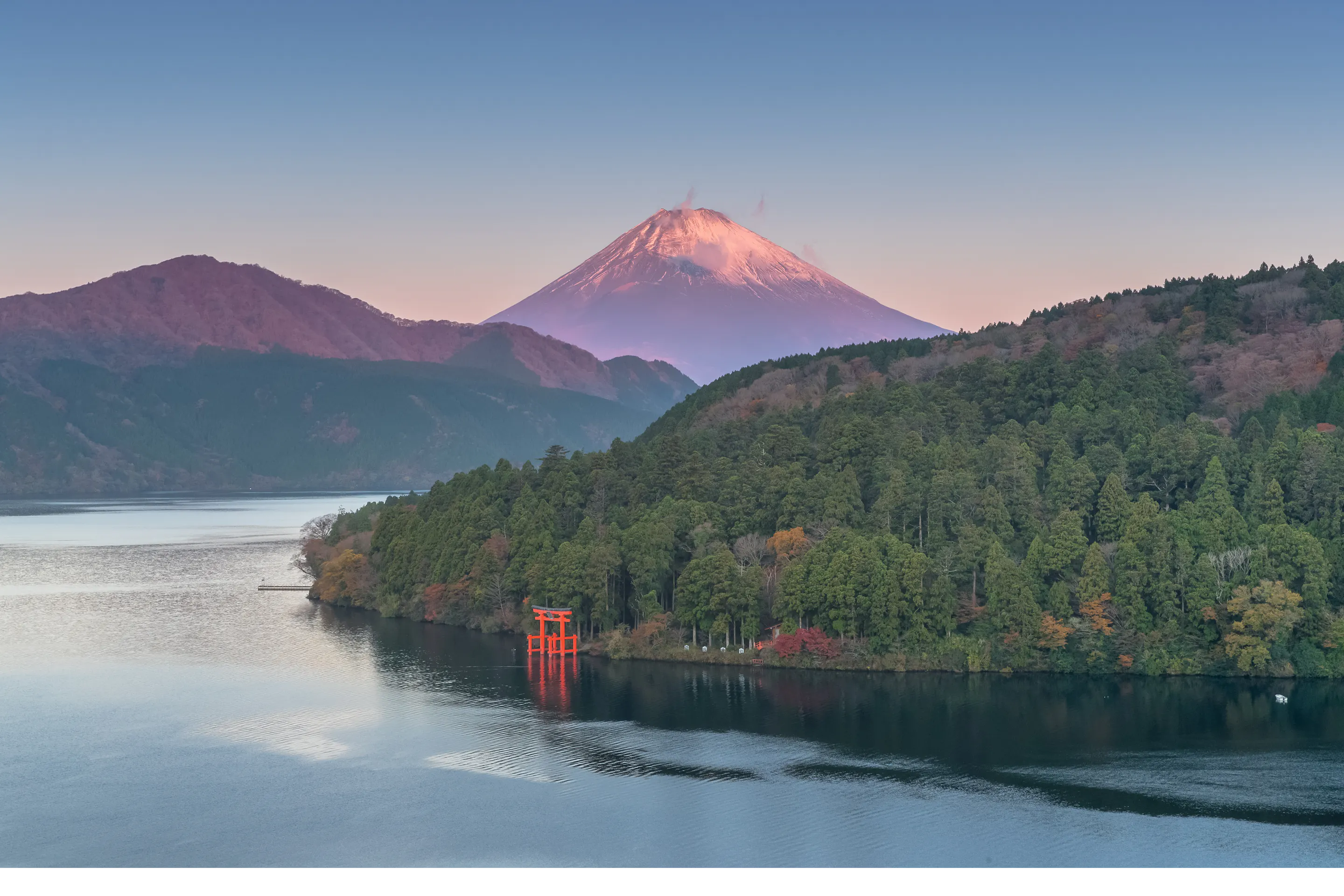Mount Fuji rises beyond Lake Ashi at sunrise, with a red torii near the shore and forested hills in autumn.