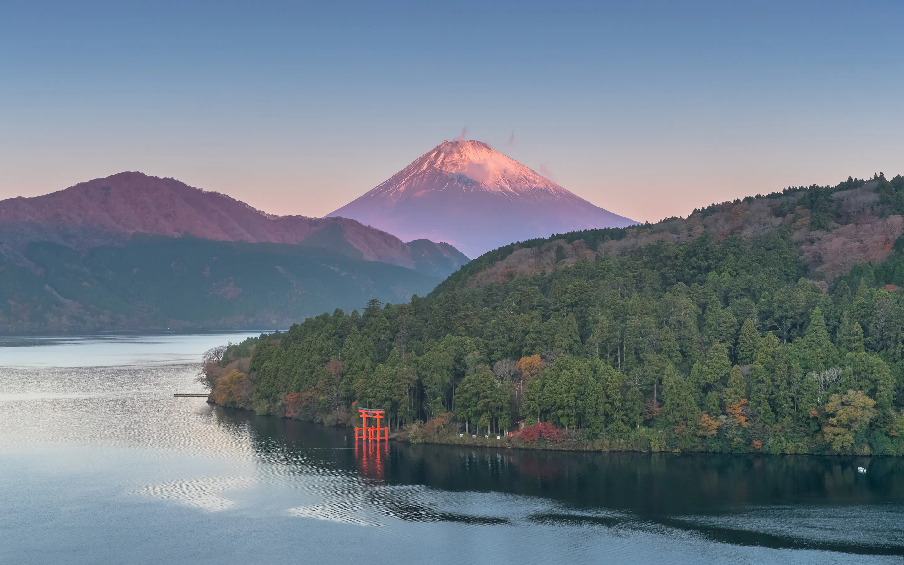 Mount Fuji rises beyond Lake Ashi at sunrise, with a red torii near the shore and forested hills in autumn.