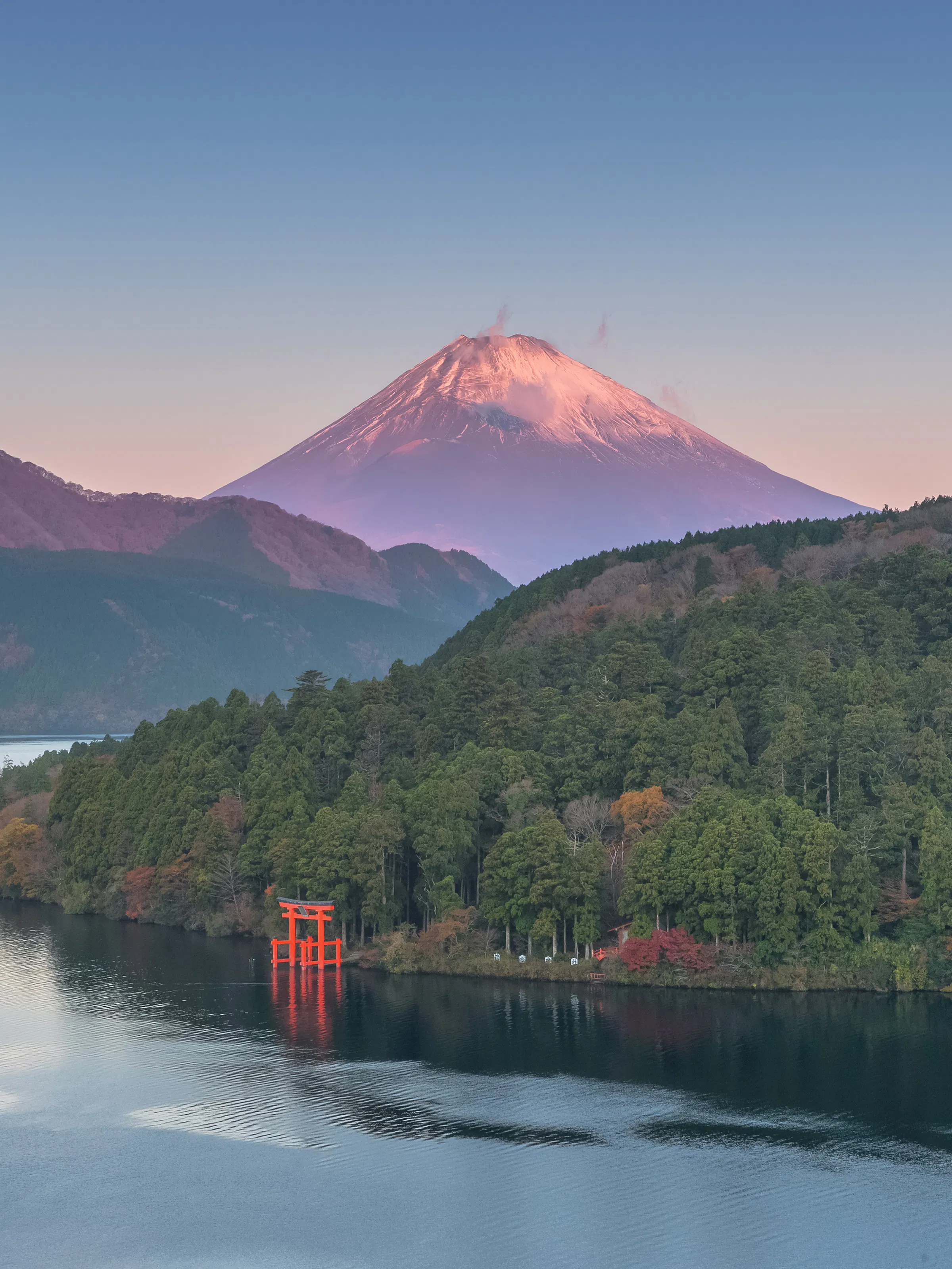 Mount Fuji rises beyond Lake Ashi at sunrise, with a red torii near the shore and forested hills in autumn.