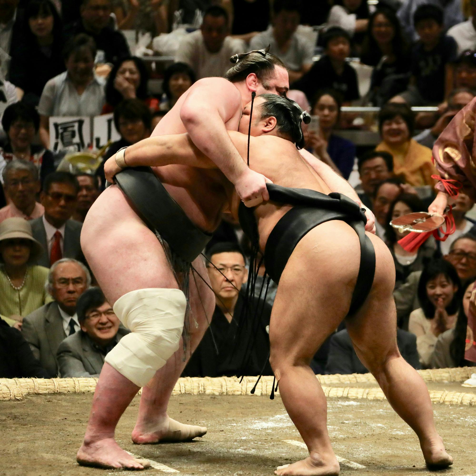 Two sumo wrestlers grapple in a ring while a referee watches, with spectators filling the arena behind them.